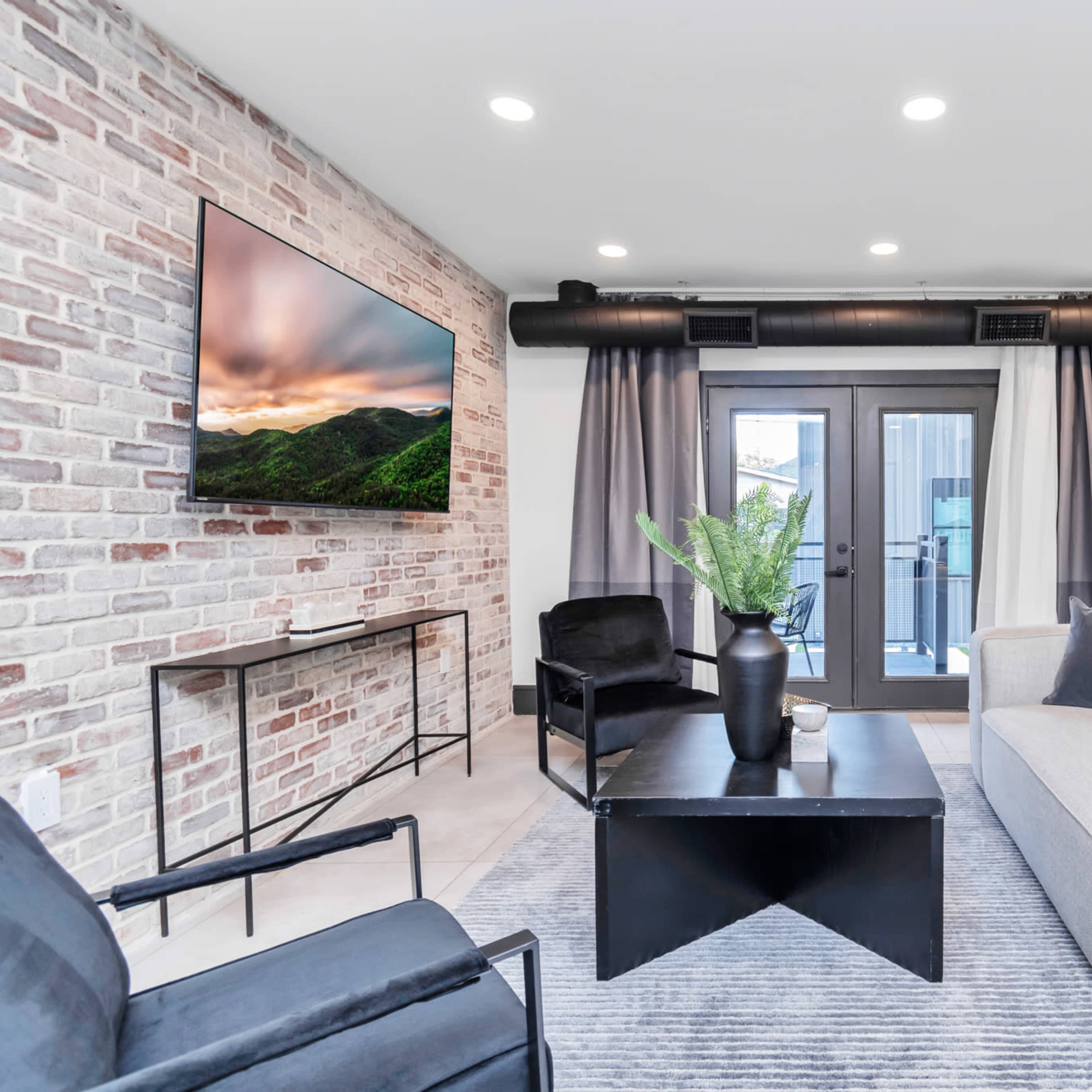 A modern living room with a gray sofa, two black chairs, a coffee table, and a large brick wall with a mounted television.
