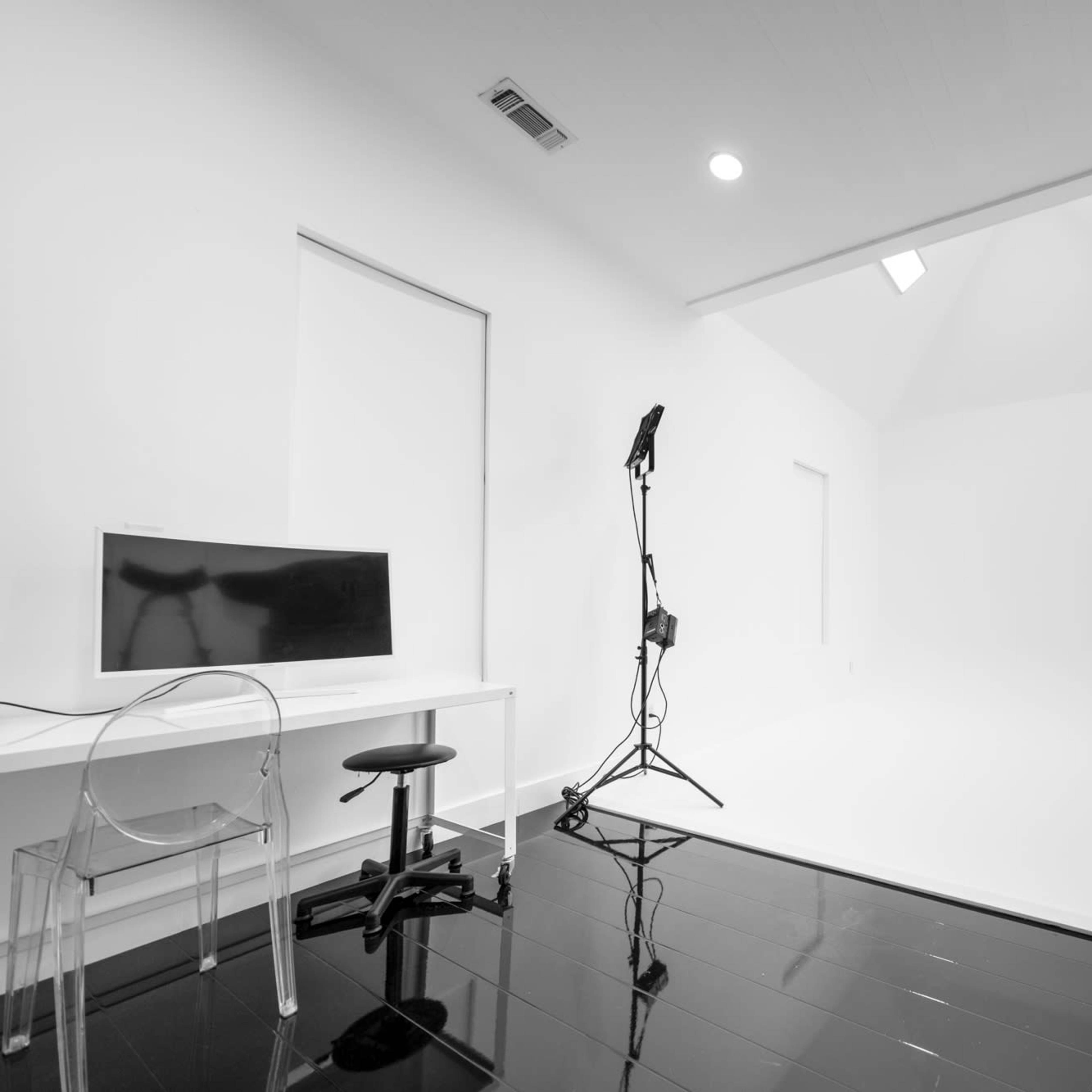 A minimalist photography studio with a white backdrop, a computer monitor on a table, and lighting equipment set up.