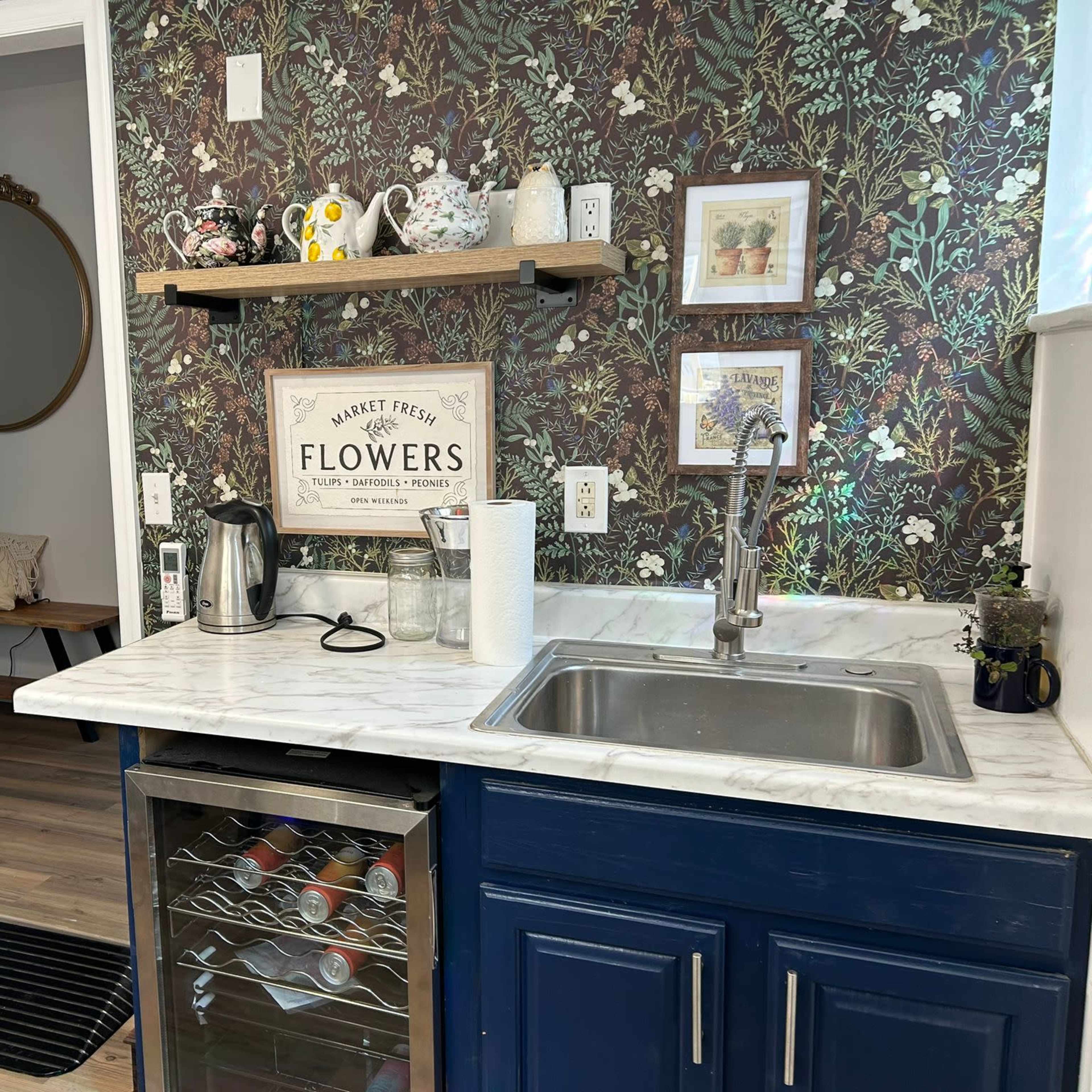 A kitchen corner with a marble countertop, a stainless steel sink, a blue cabinet with a visible wine cooler, and decorative wall elements featuring floral wallpaper and various kitchen items.