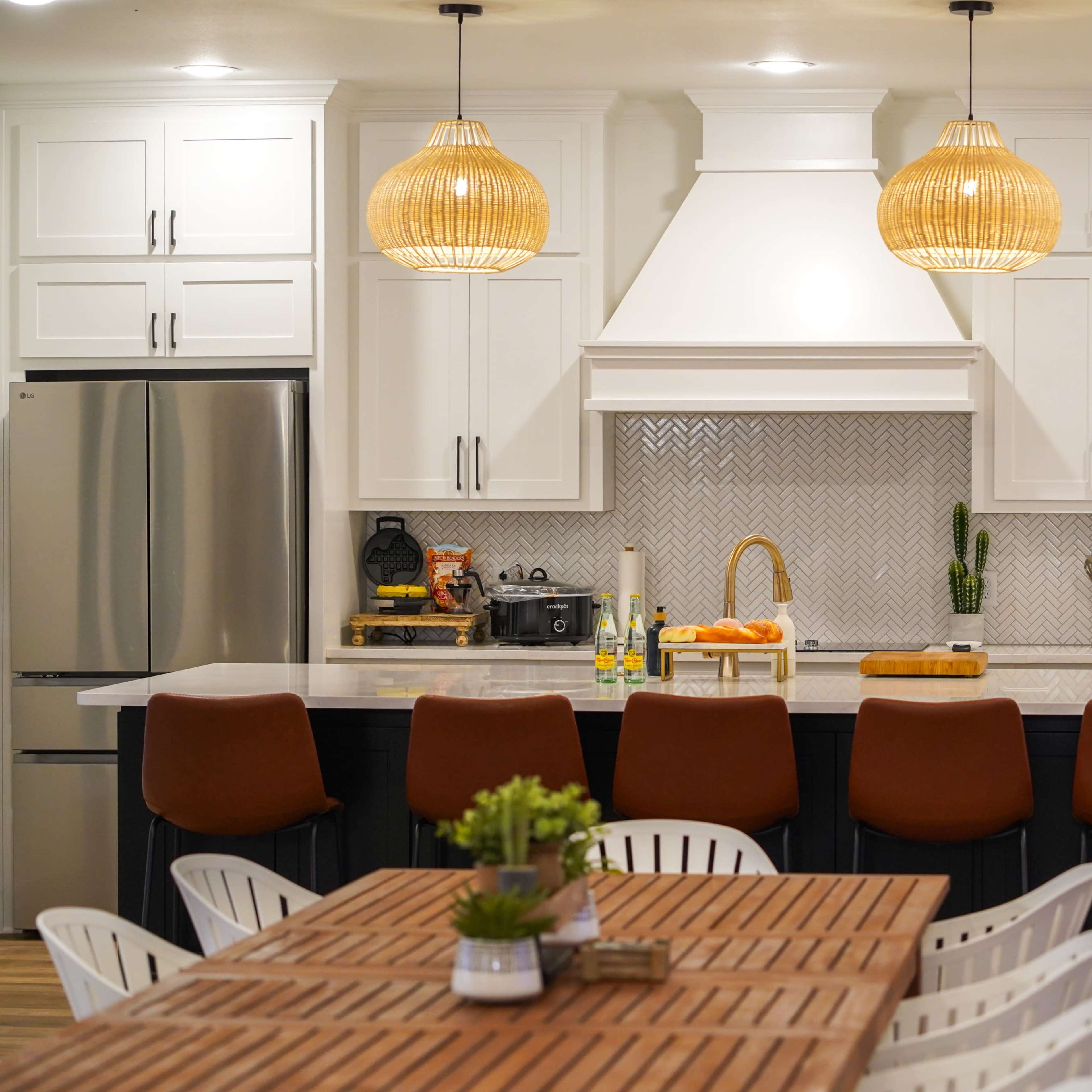A modern kitchen with white cabinetry, a large island with bar stools, and an adjacent dining table, all featuring a neutral color palette and pendant lighting.