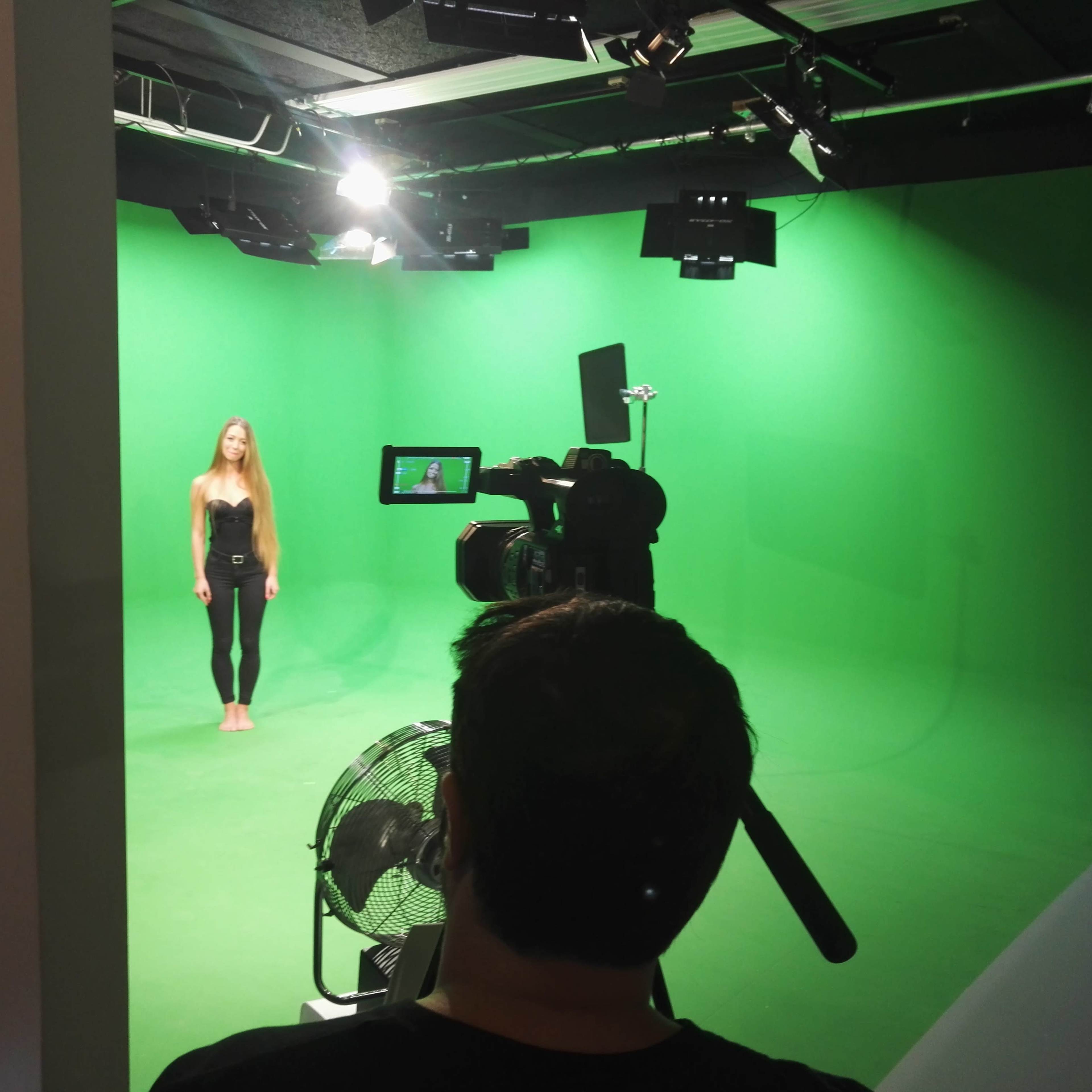 A person stands barefoot in front of a green screen, while a camera is positioned to capture the scene.