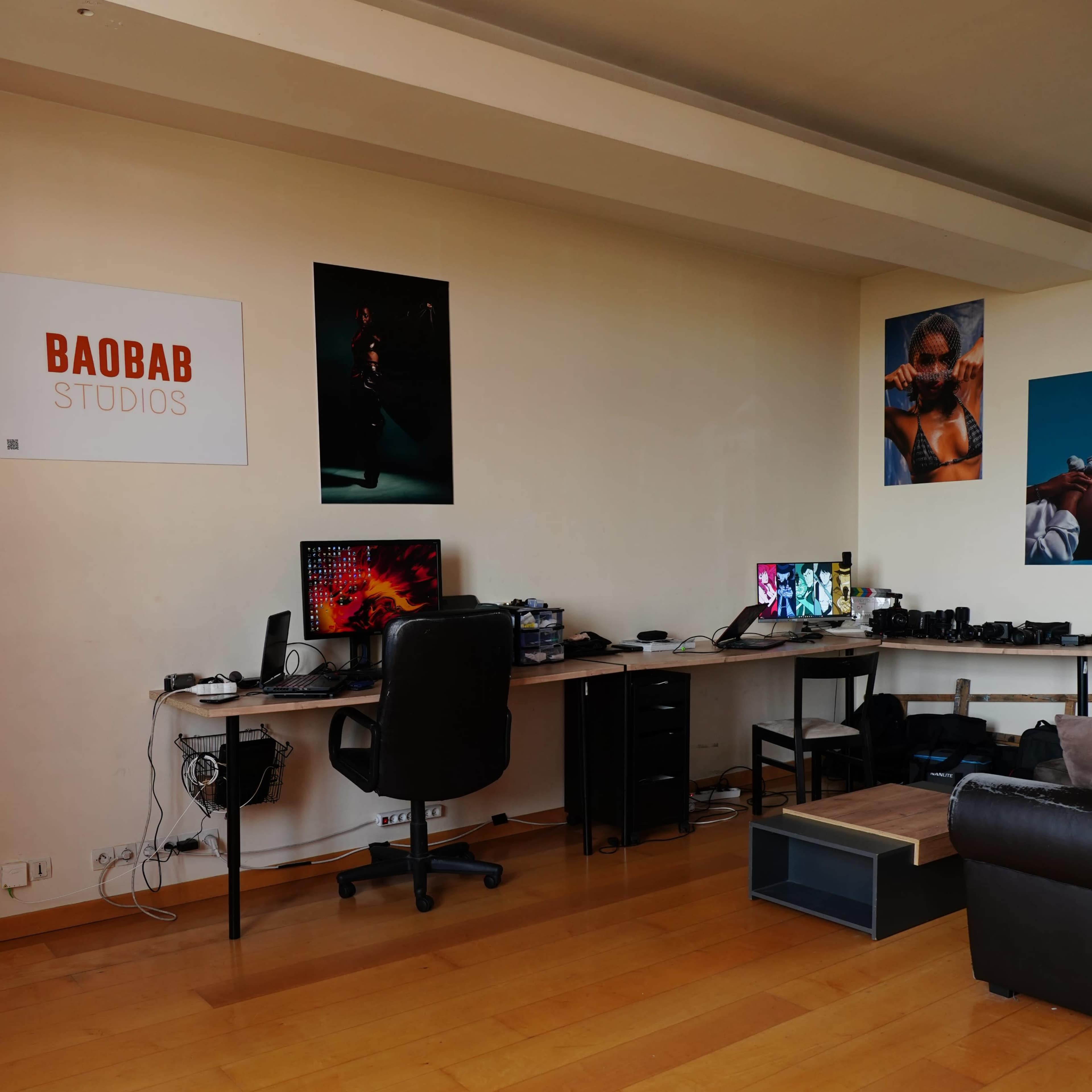 A studio workspace with two desks, computers, and various equipment against a wall adorned with posters.