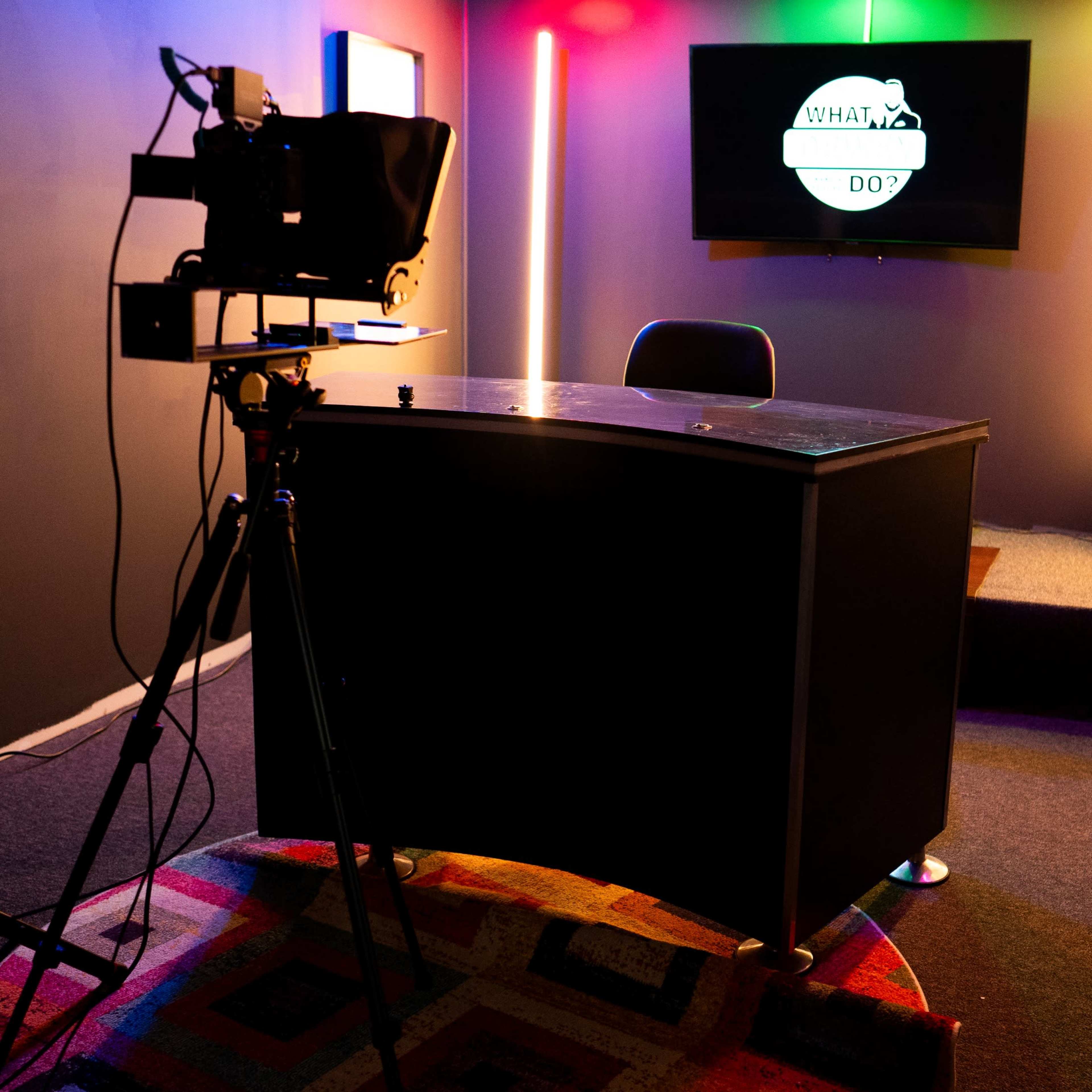 A desk and chair are set up in a small room equipped with colorful lights and a television screen displaying a logo.