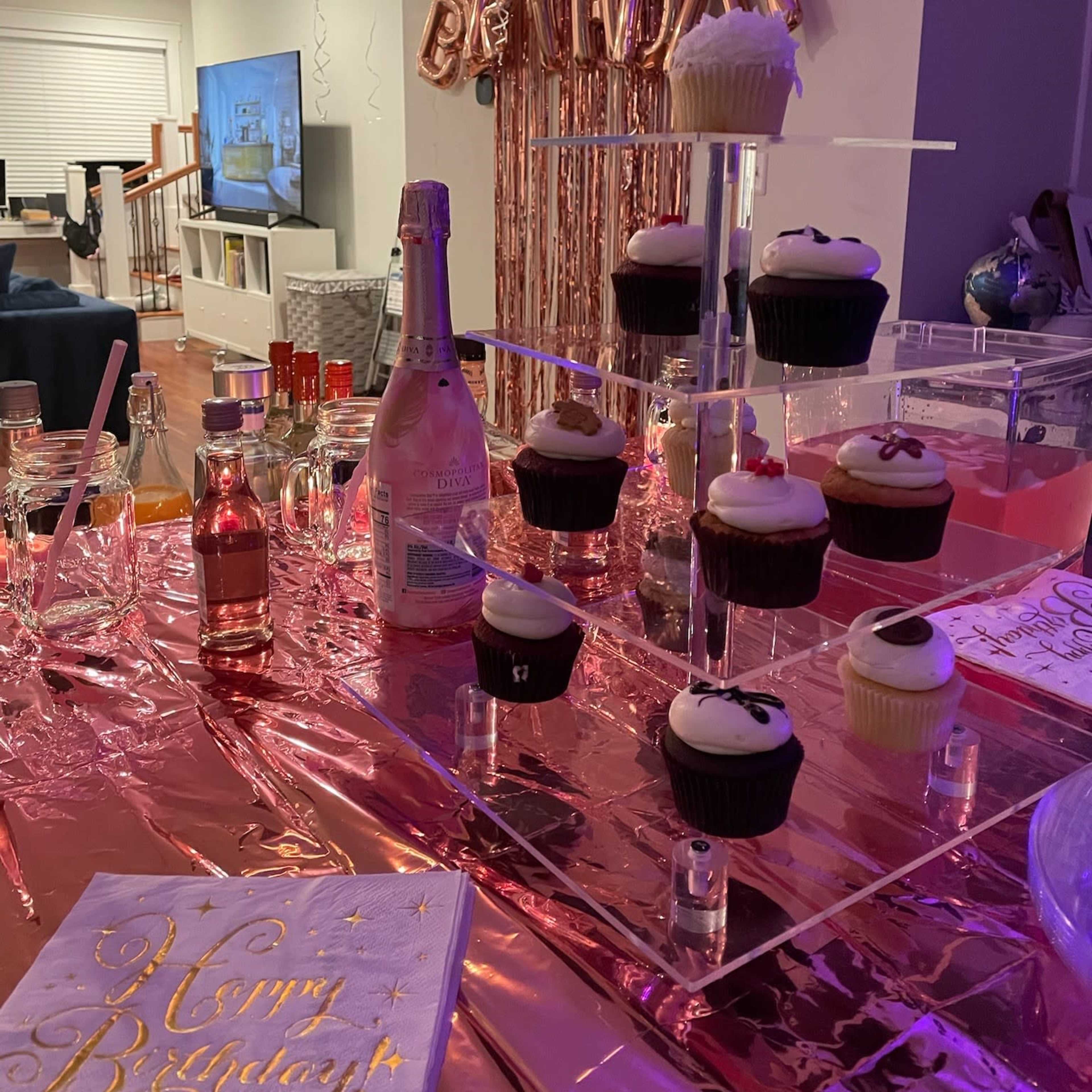 A birthday celebration setup with cupcakes on a clear stand, a bottle of sparkling beverage, and a "Happy Birthday" decoration in the background.