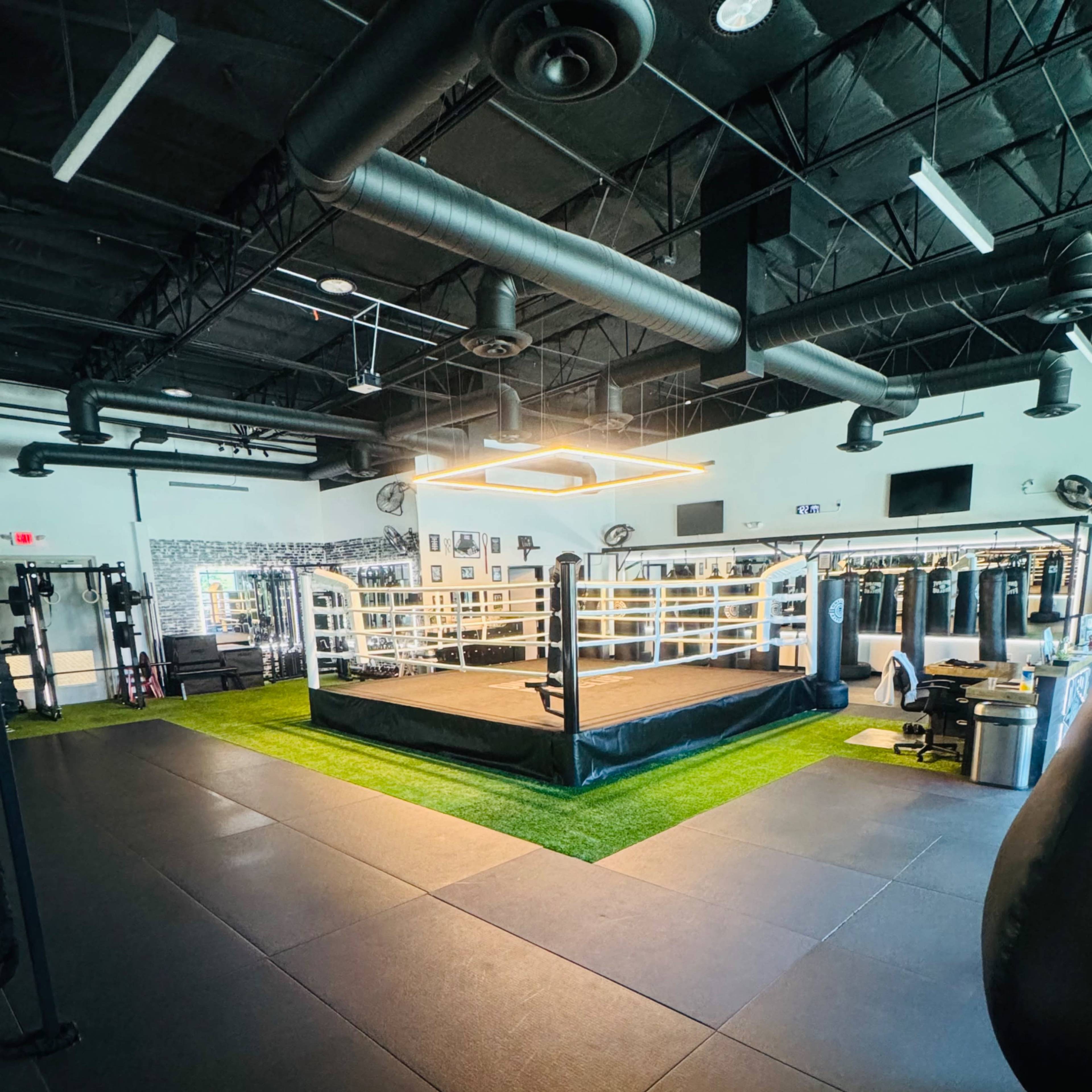 A spacious boxing gym featuring a central ring surrounded by exercise equipment and padded flooring.