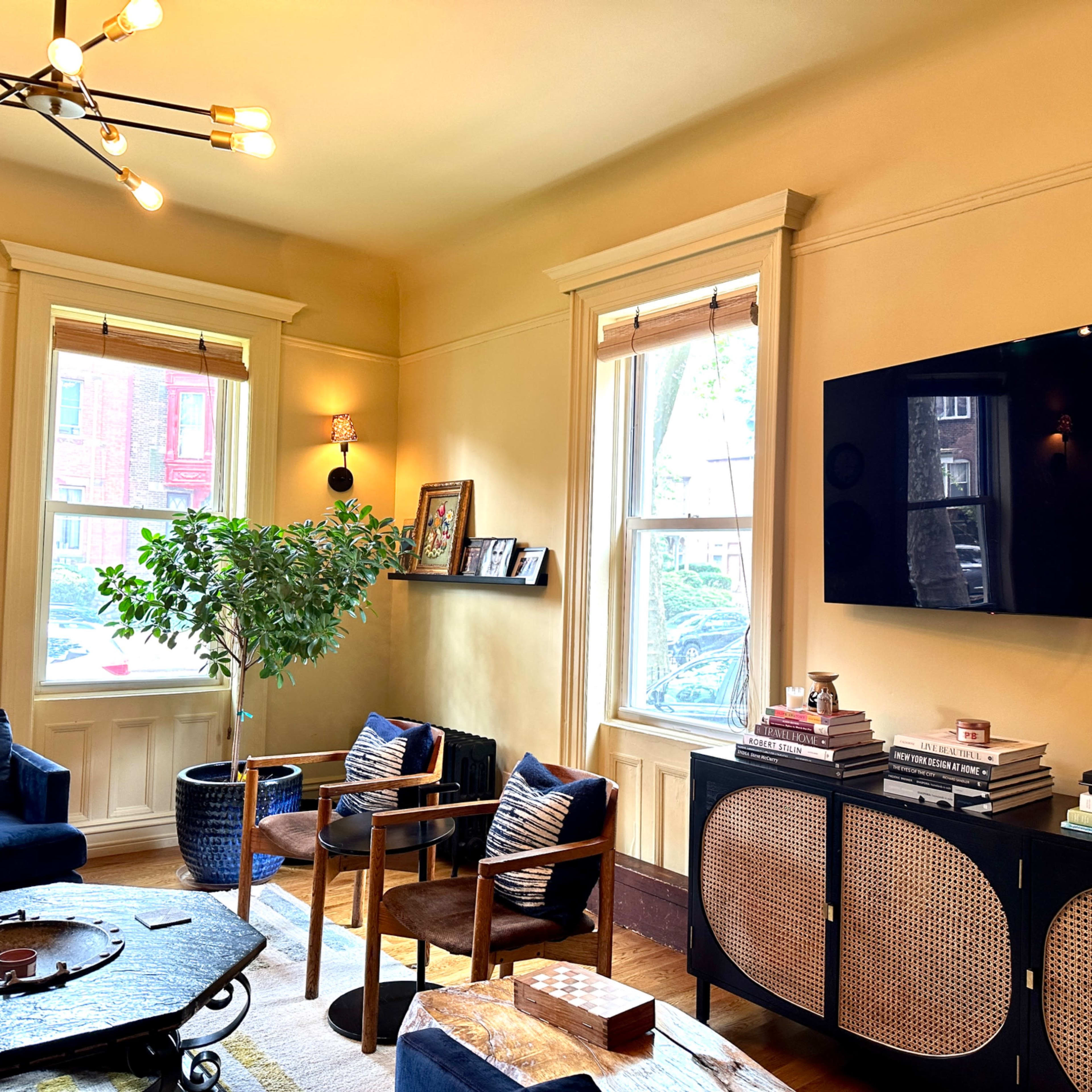 A cozy living room featuring a television mounted on a wall, a dark cabinet with decorative books, a potted plant, and a seating area with blue chairs and a circular table.