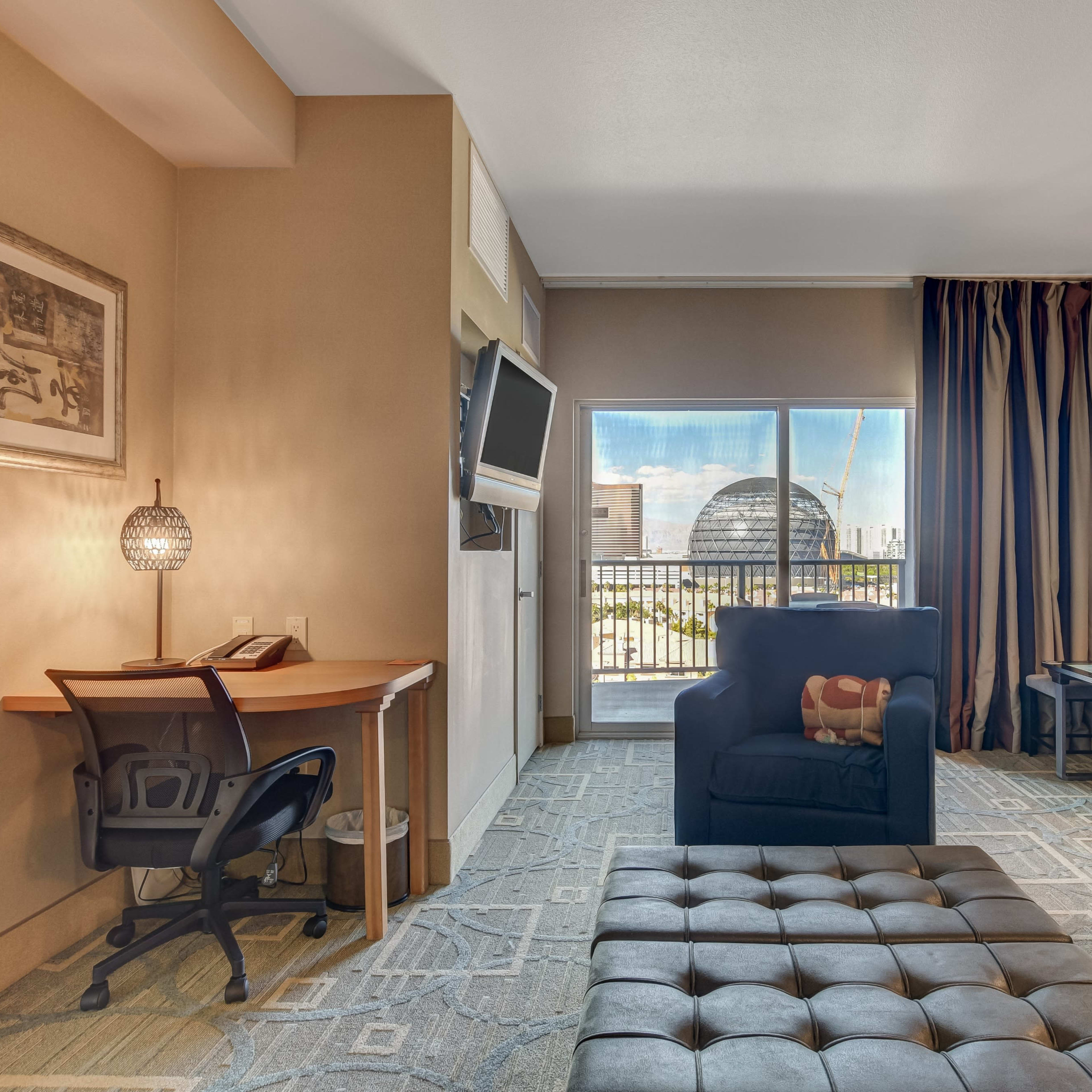 A well-furnished hotel room features a desk, a plush seating area, and a large window overlooking a cityscape.