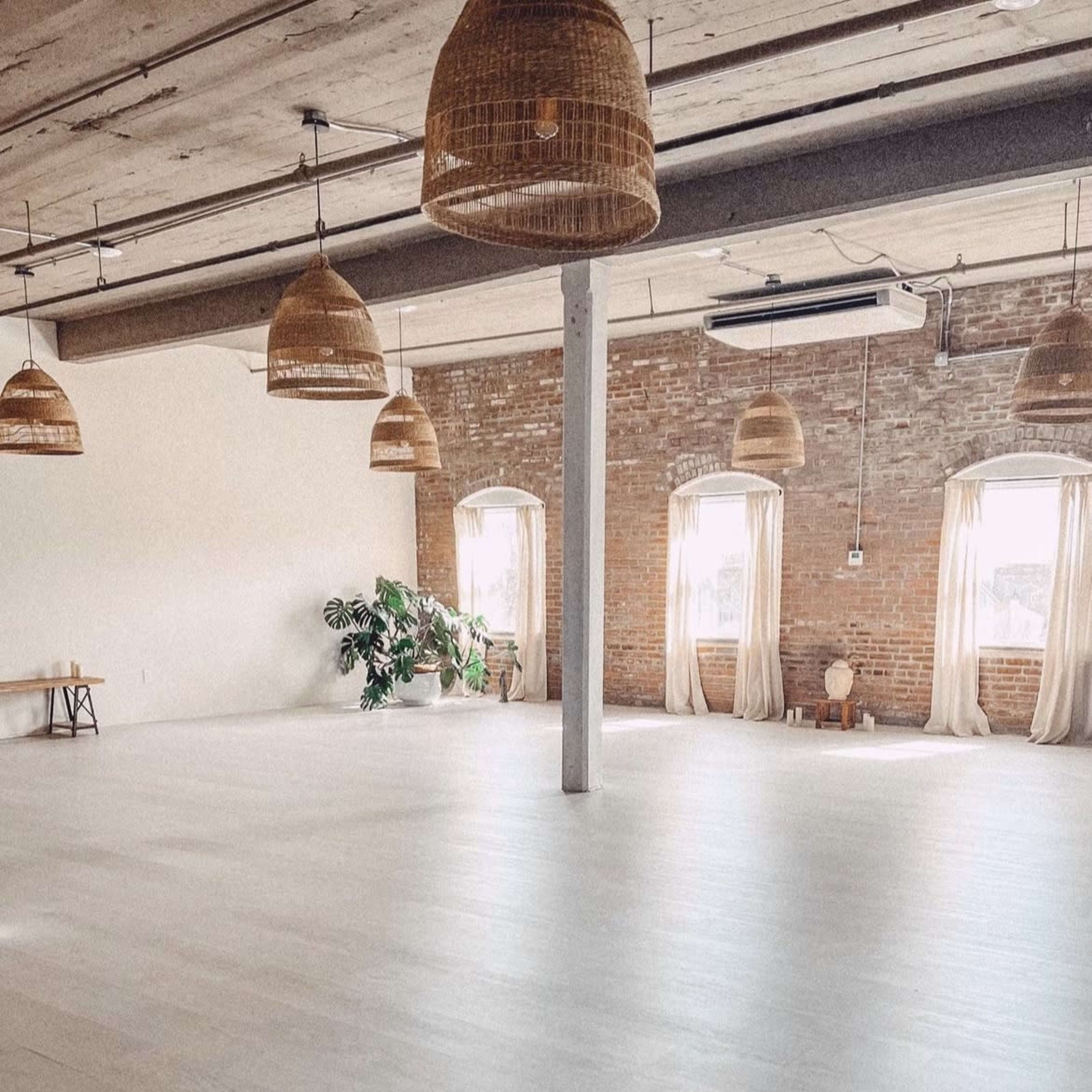 A spacious, lightly furnished room with wooden floors, large windows, and hanging wicker light fixtures.