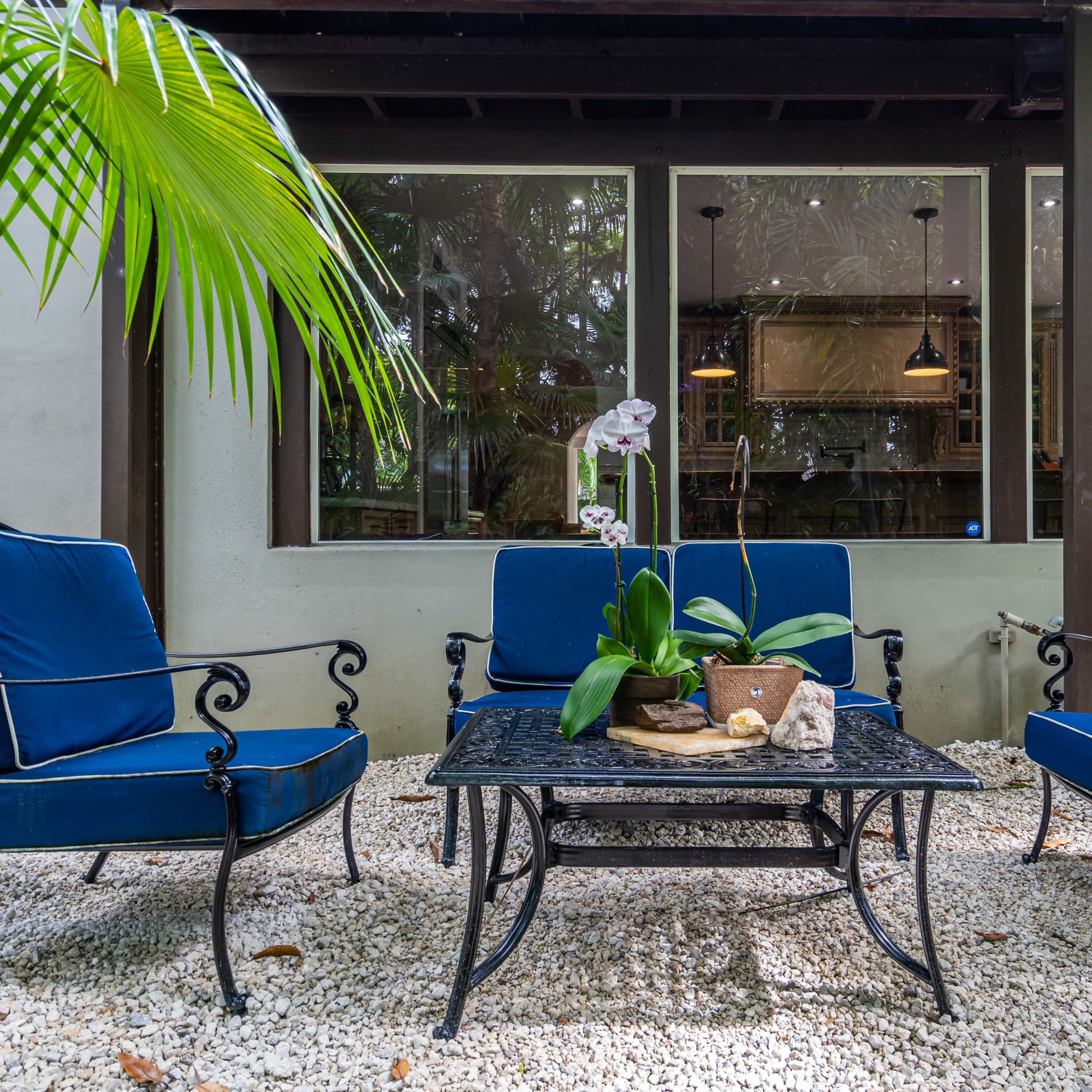 A patio area features four blue cushioned chairs surrounding a black metal coffee table, set on a bed of white gravel with greenery and large windows in the background.