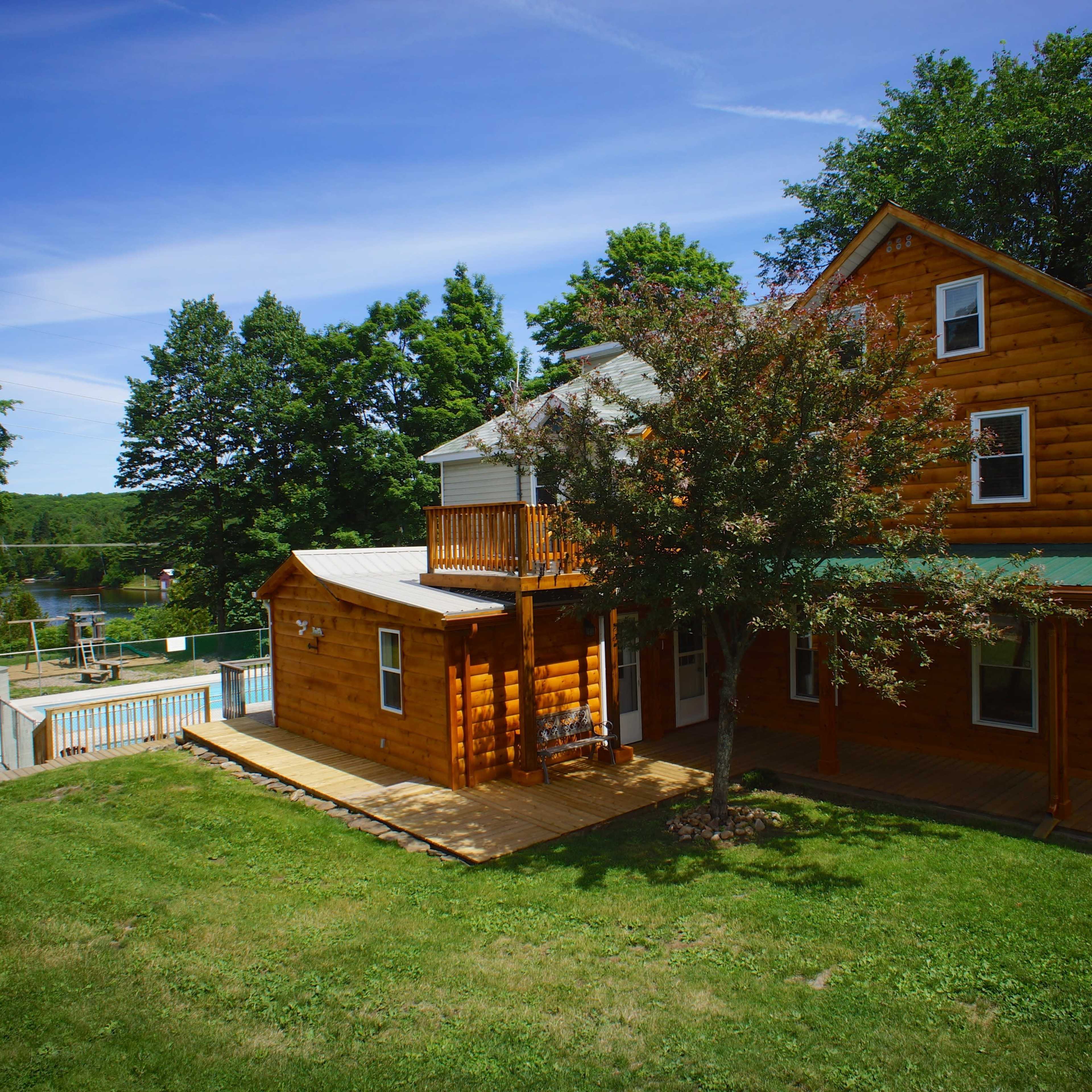 A large wooden house with a green roof and a deck overlooks a pool and a lake surrounded by trees.