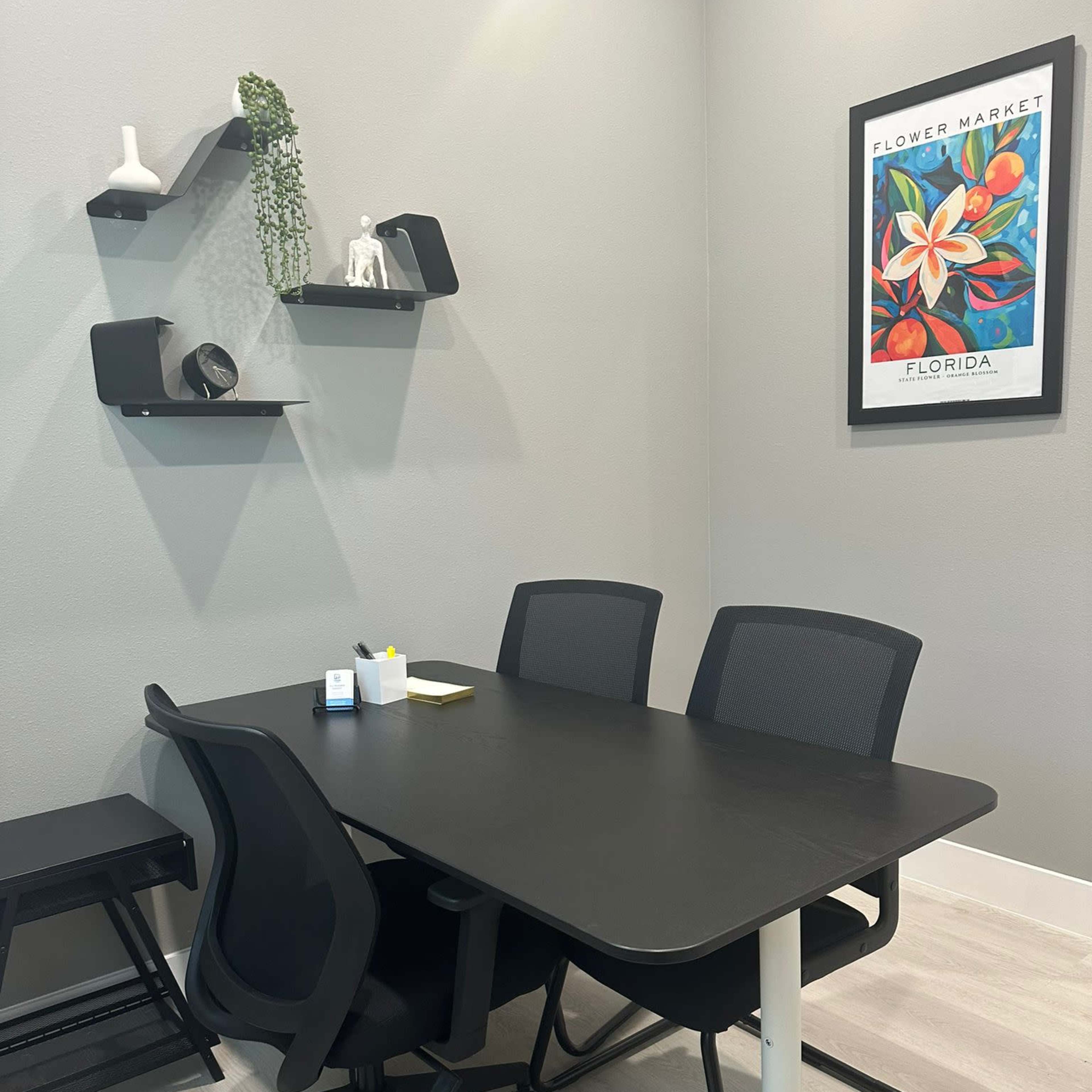 A modern conference room features a black table with three ergonomic chairs, a wall shelf displaying decorative items, and a framed artwork.
