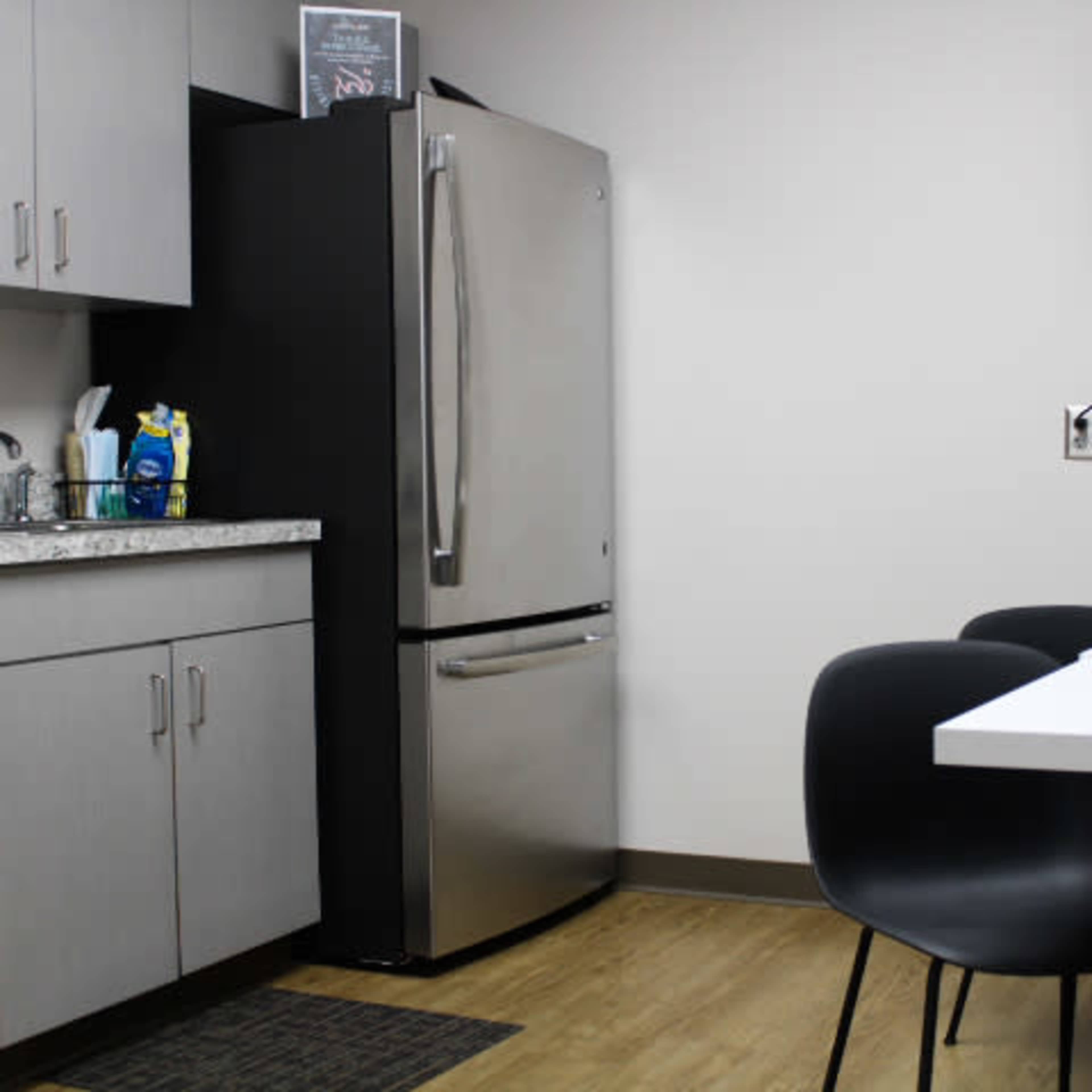 A modern kitchen area featuring a stainless steel refrigerator, a sink with countertop space, a dining table with four black chairs, and a wall-mounted television.