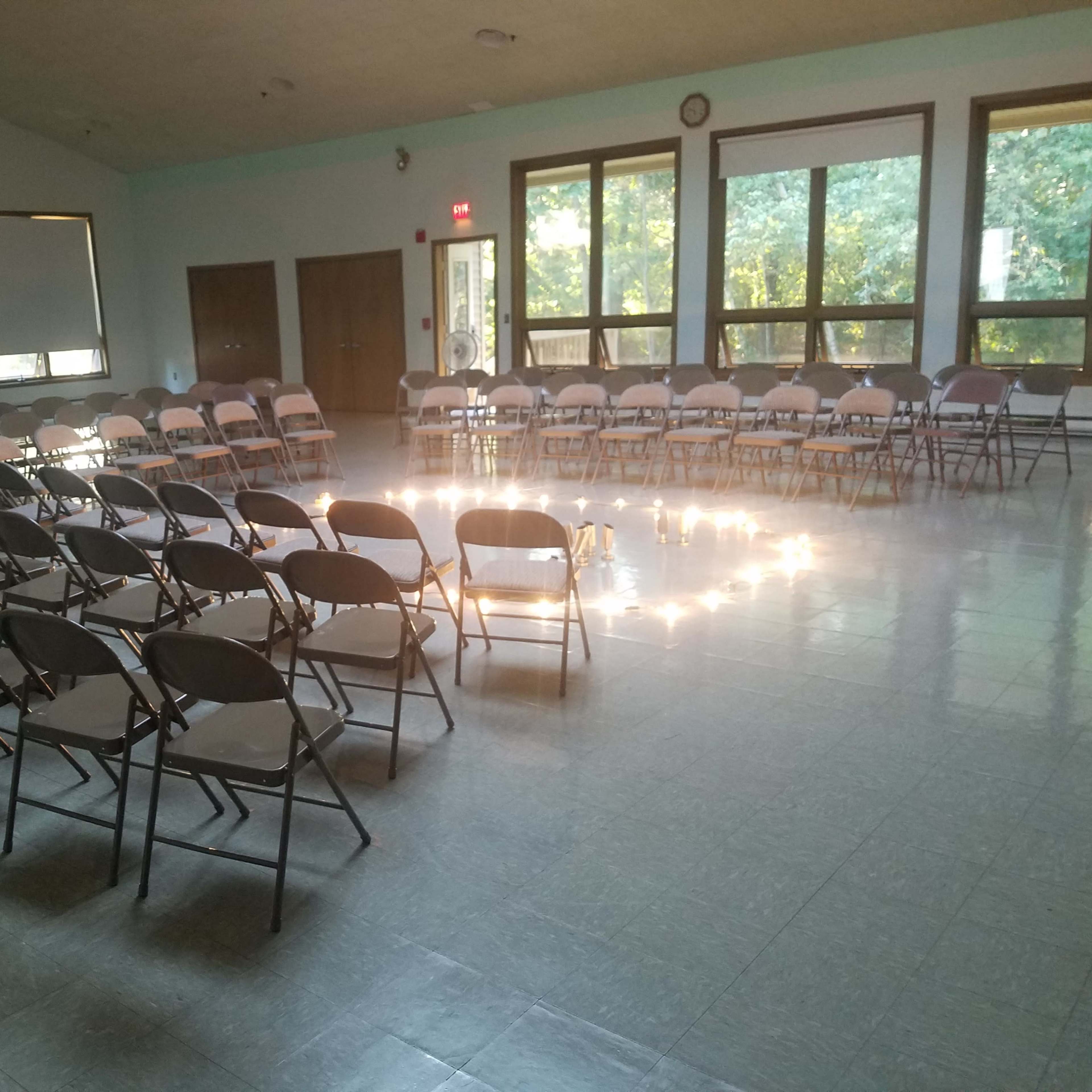 A room is arranged with rows of folding chairs facing a circular formation in the center, illuminated by natural light from large windows.