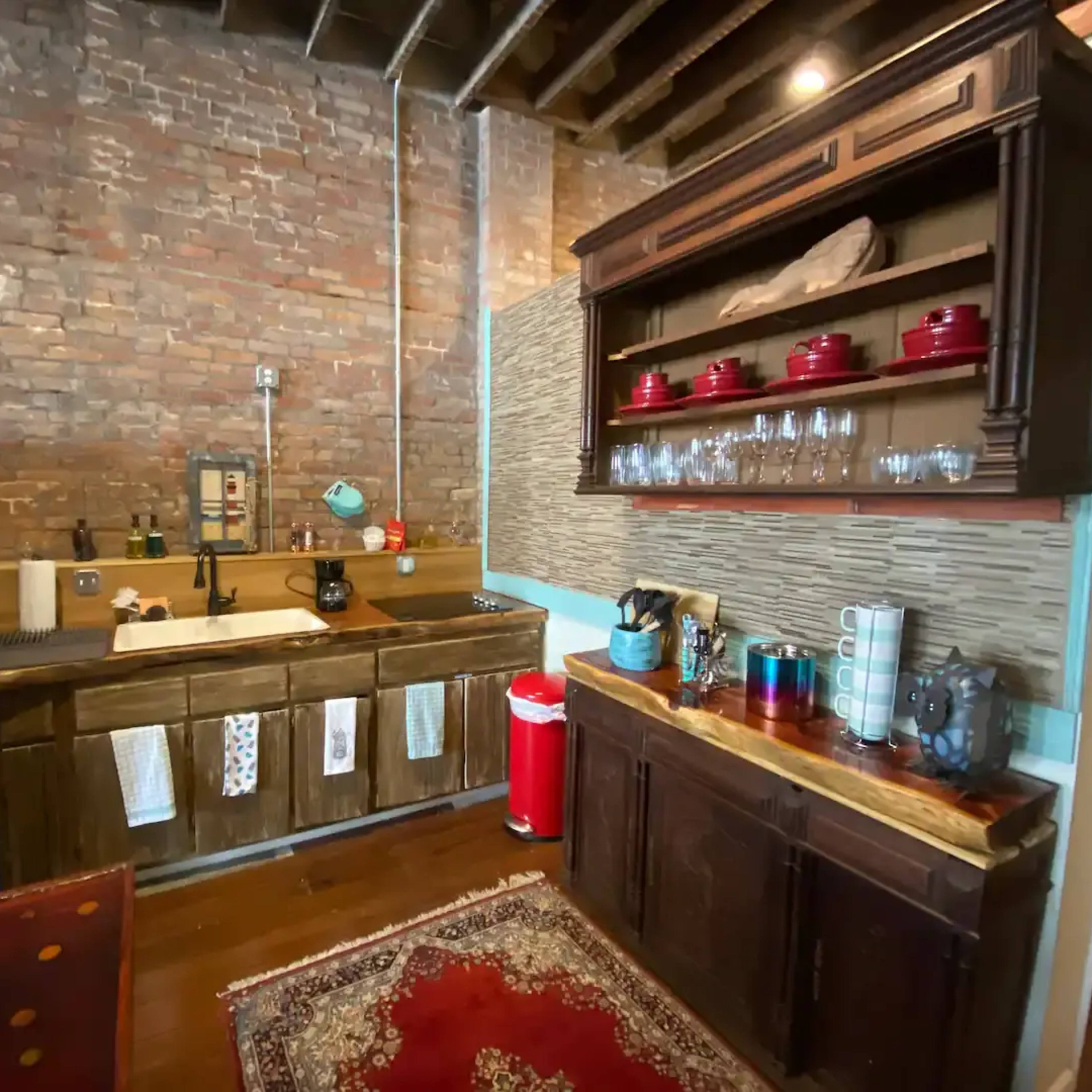 A rustic kitchen with exposed brick walls, wooden cabinetry, a farmhouse sink, and shelves displaying red dishware and glassware.