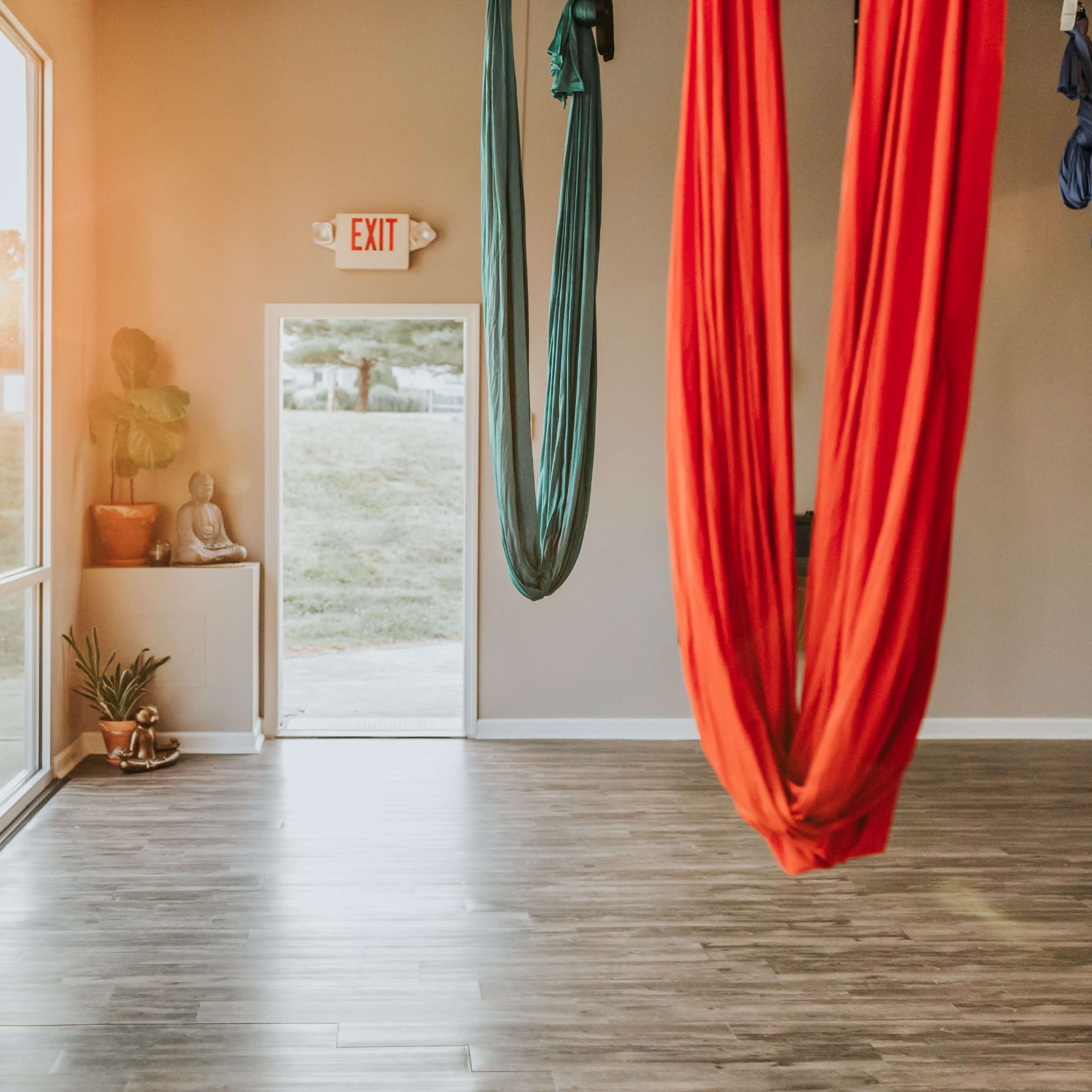 An aerial yoga studio with red and blue fabric hammocks hanging from the ceiling, and sunlight streaming through large windows.
