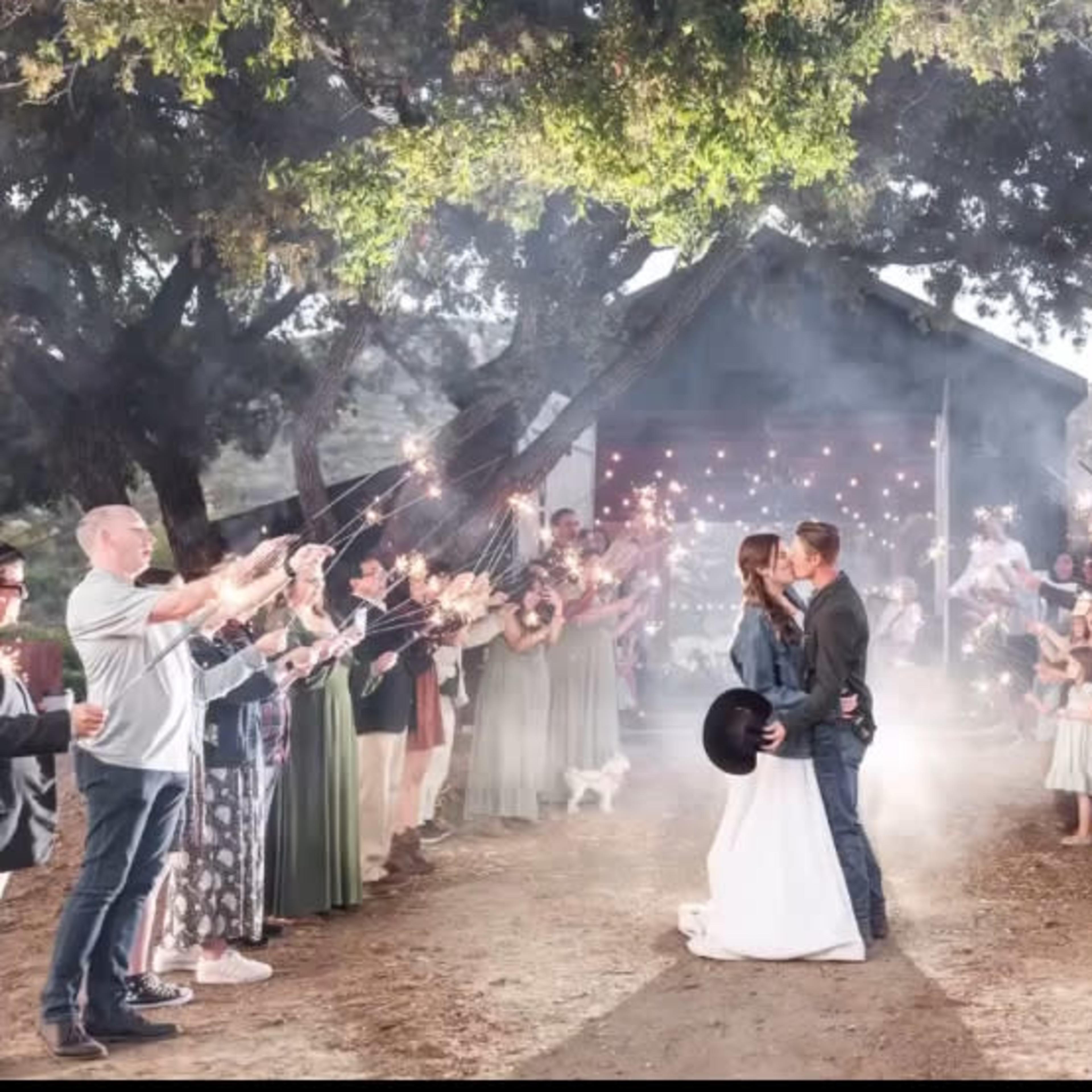 A couple kisses in front of a rustic building as guests hold sparklers and celebrate their wedding.