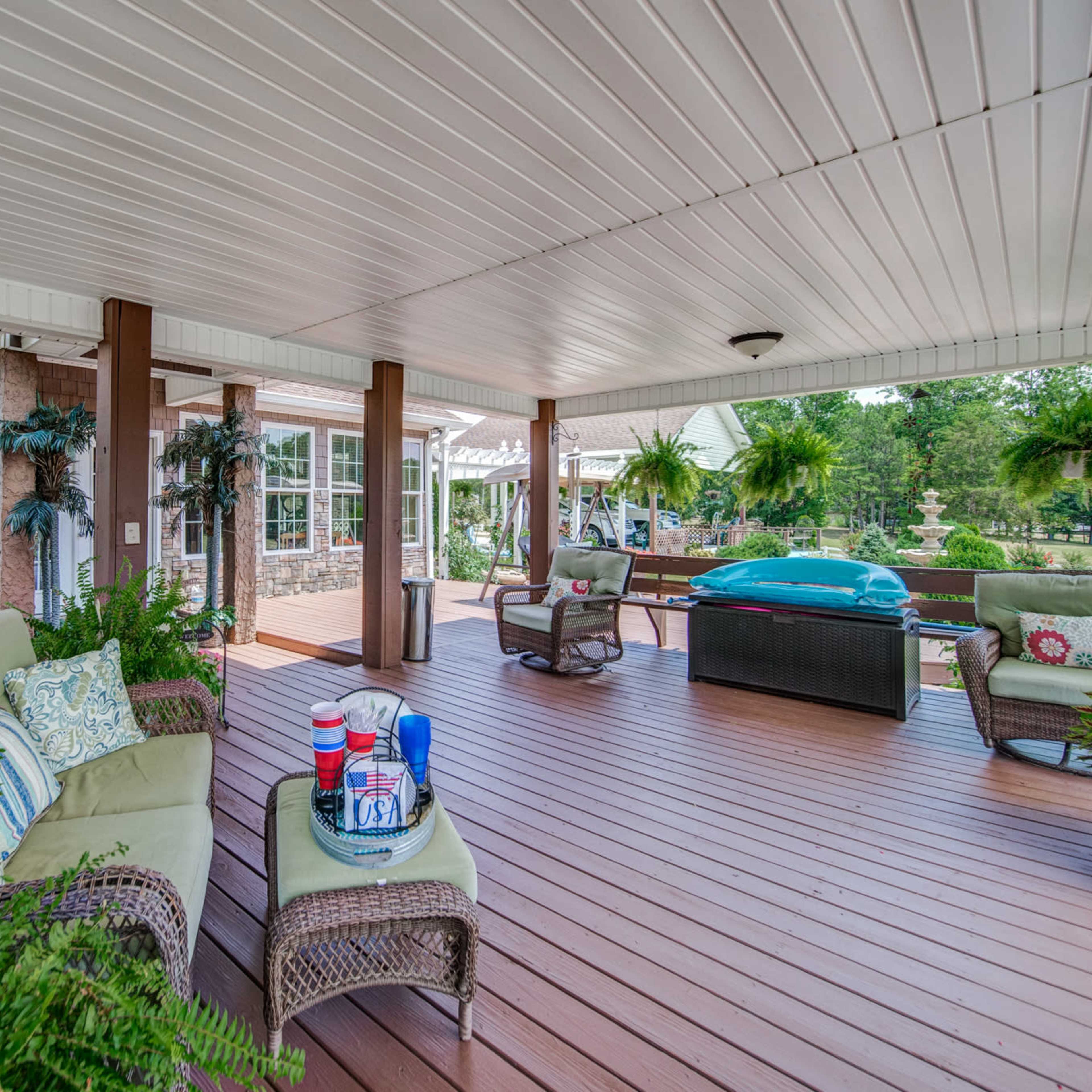 An outdoor covered patio with comfortable seating, potted plants, and a hot tub in a landscaped yard.