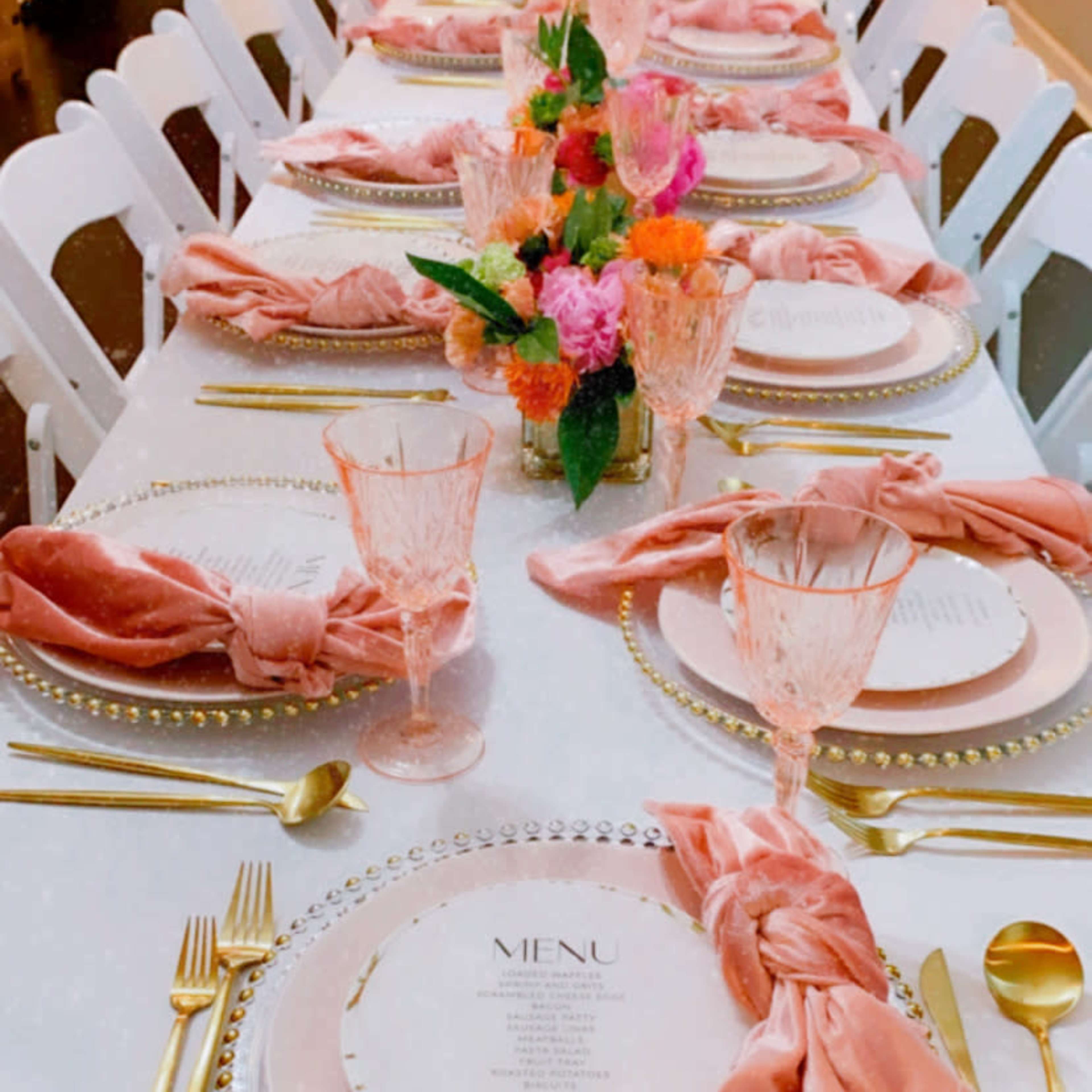A long dining table is elegantly set with gold utensils, pink napkins, and floral centerpieces.
