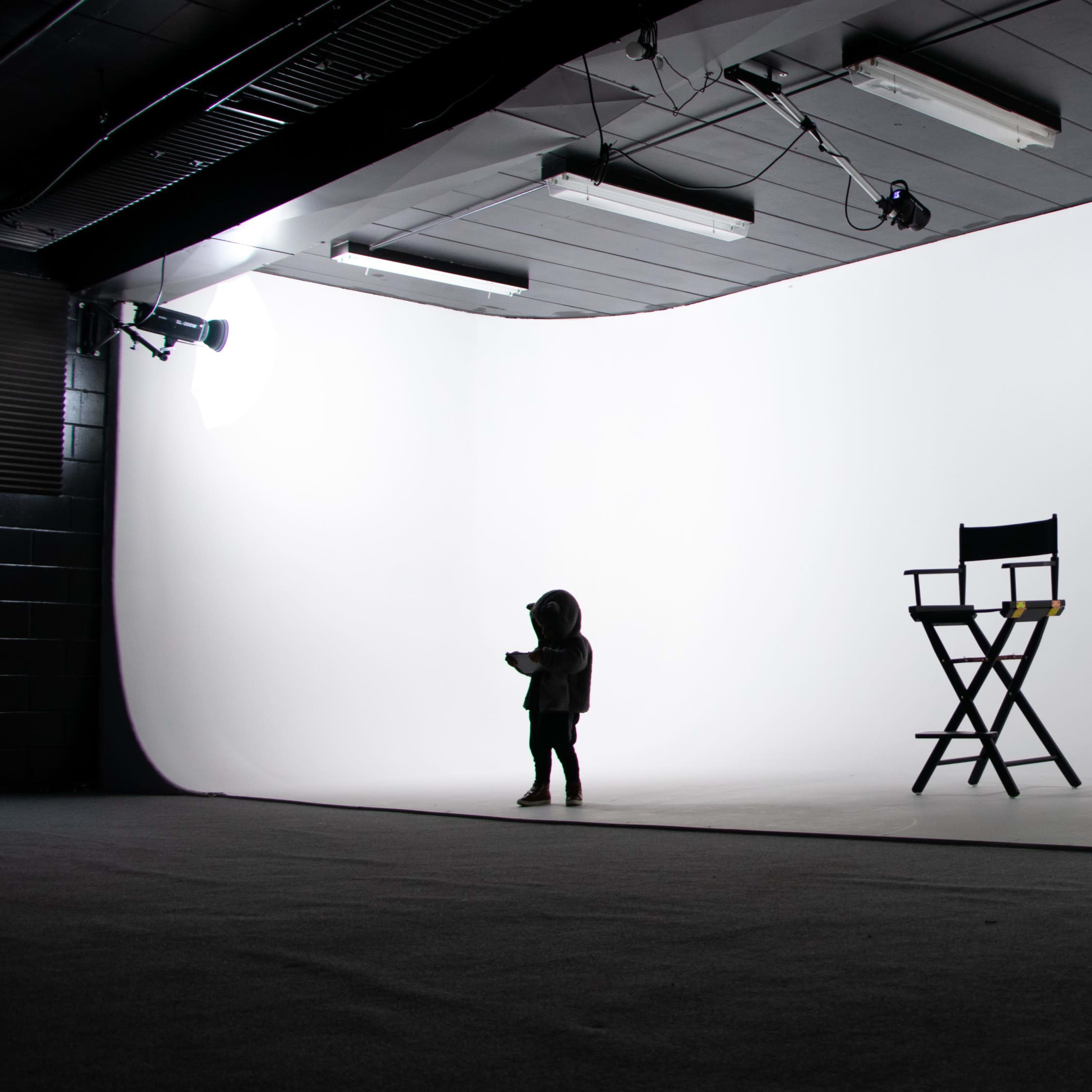 A child in silhouette stands in a spacious photography studio next to an empty director's chair against a white backdrop.