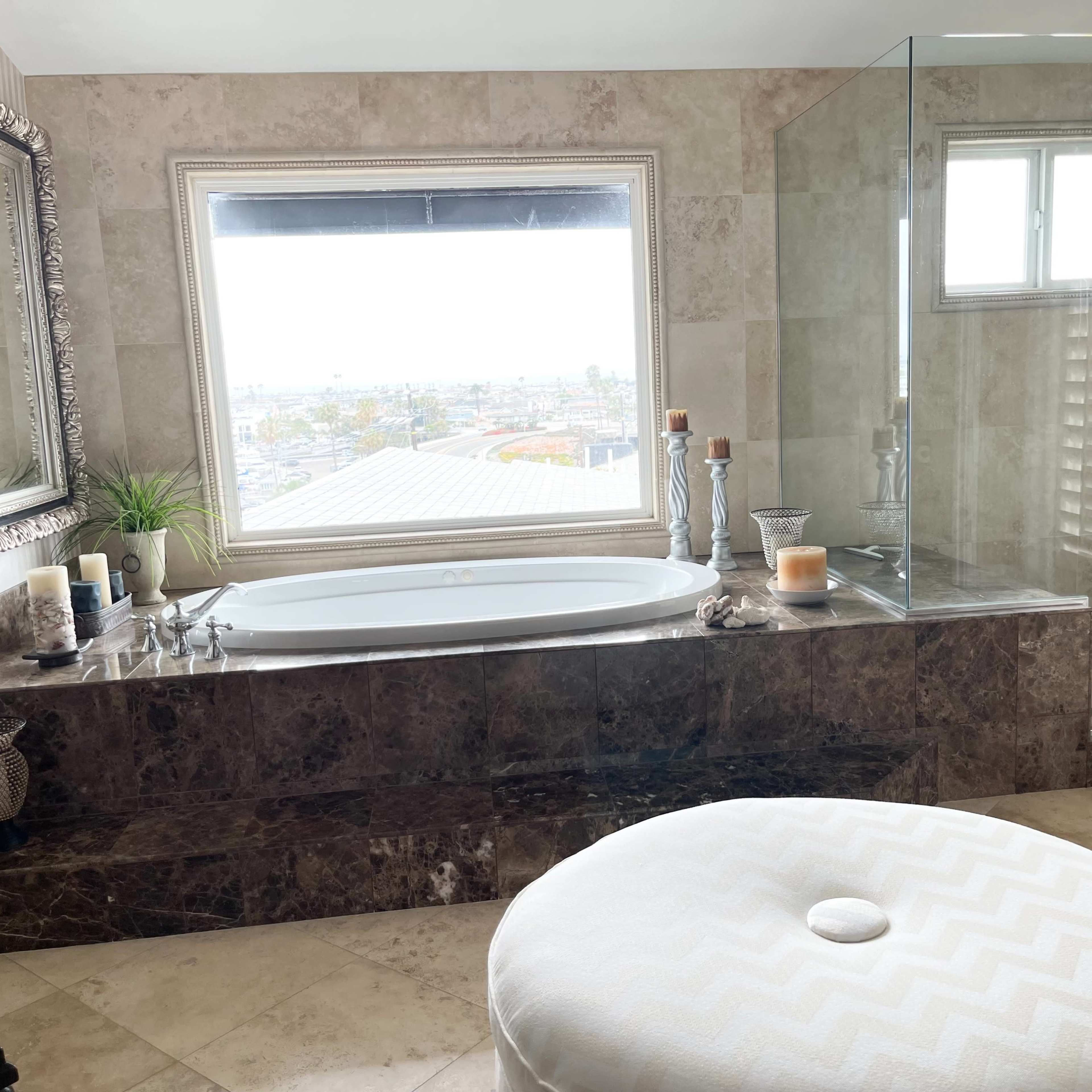 A spacious bathroom features a soaking tub beside a large window, a glass-enclosed shower, and decorative candles on a marble countertop.