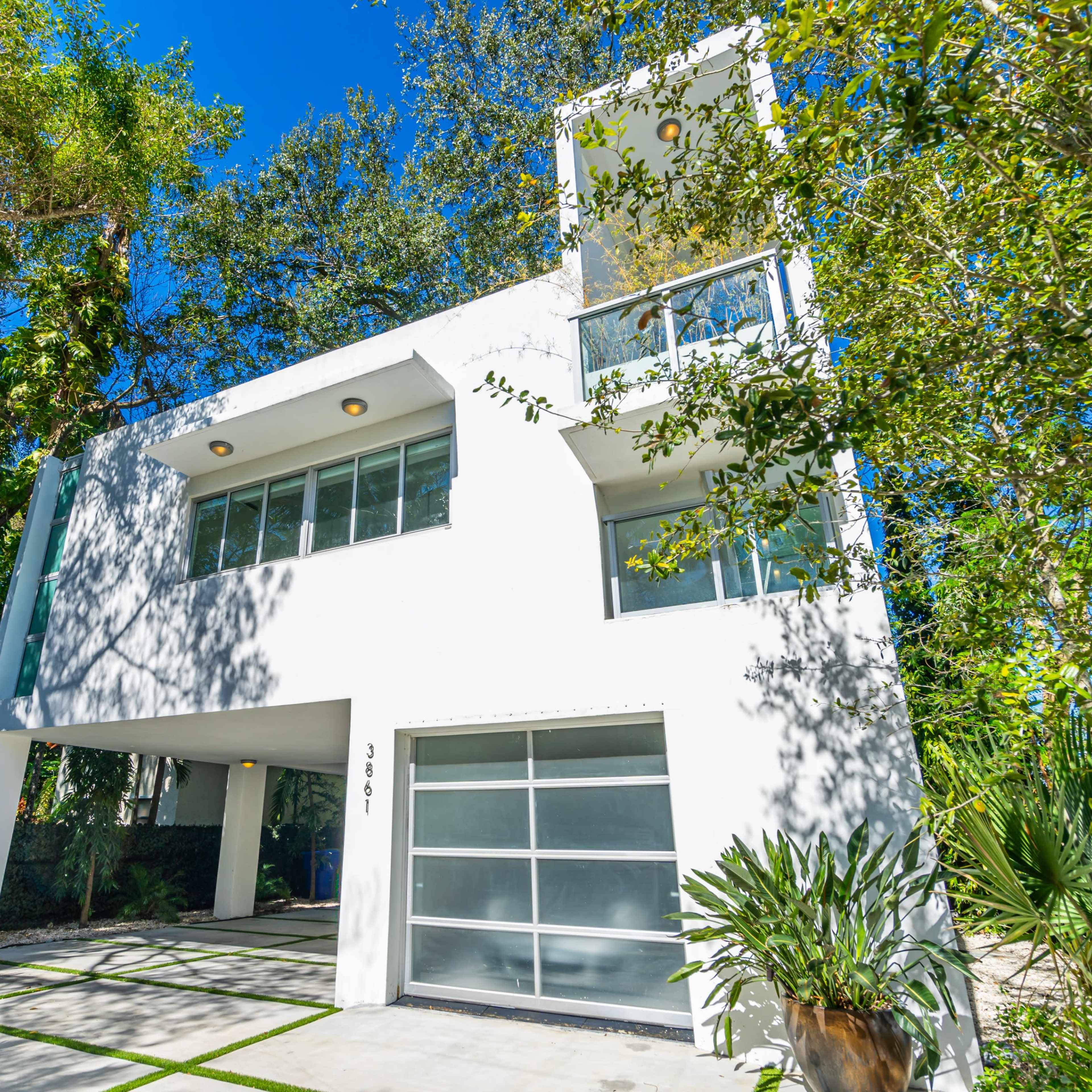 A modern white three-story house surrounded by lush greenery and palm trees.