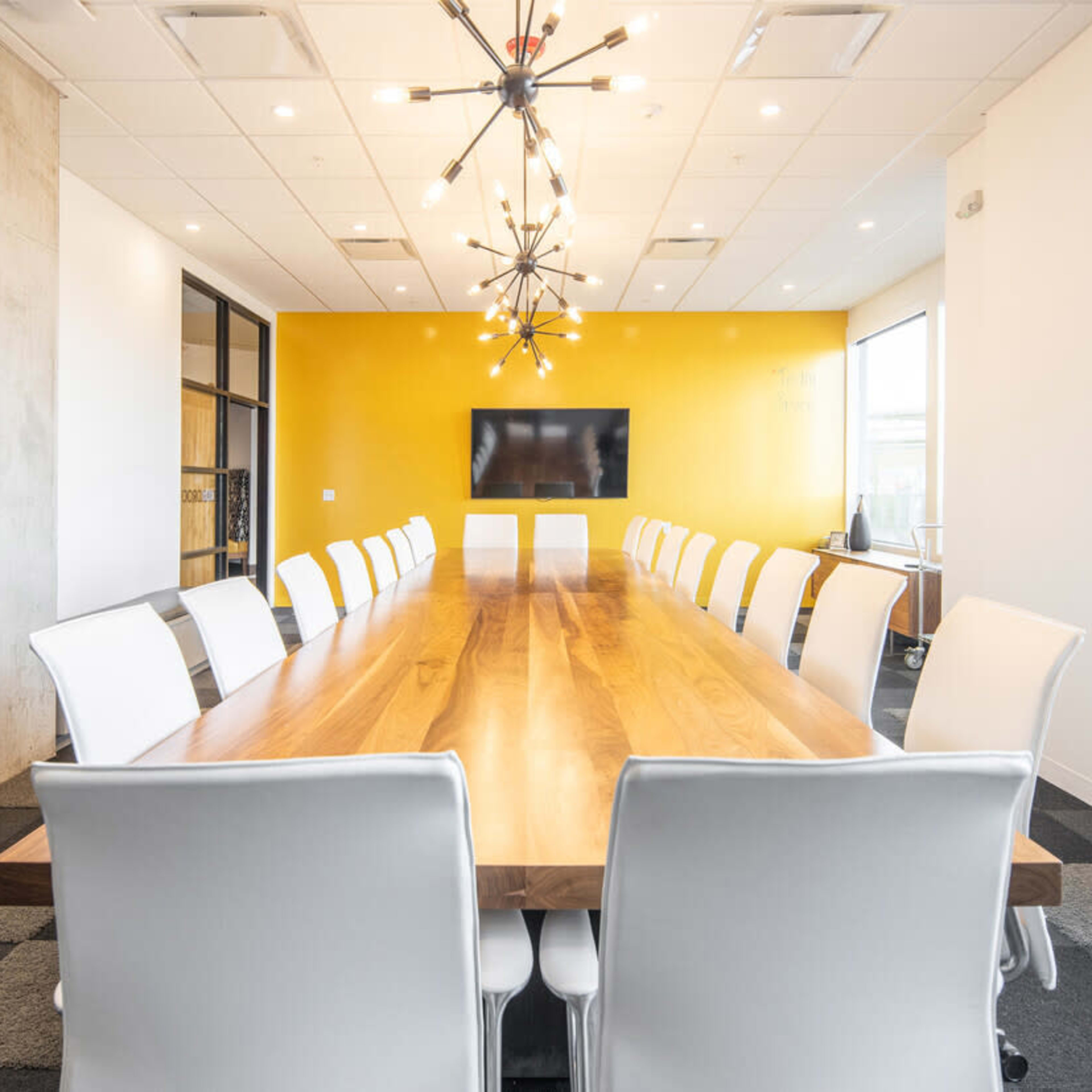 A long wooden conference table with white chairs is set in a brightly lit meeting room featuring a yellow accent wall and a modern light fixture.