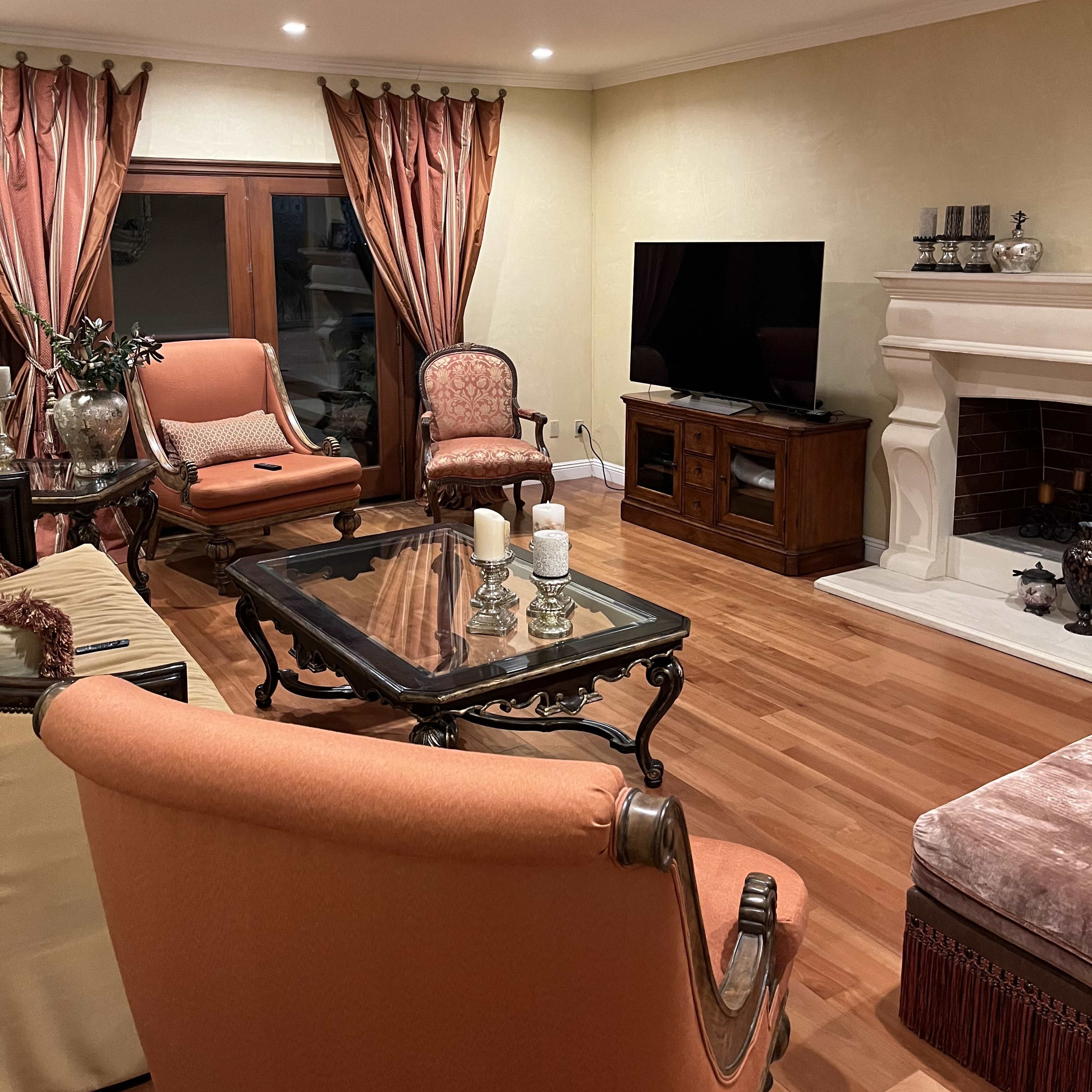 A spacious living room with wooden flooring, elegant curtains, and a fireplace, featuring a glass coffee table and a large television.
