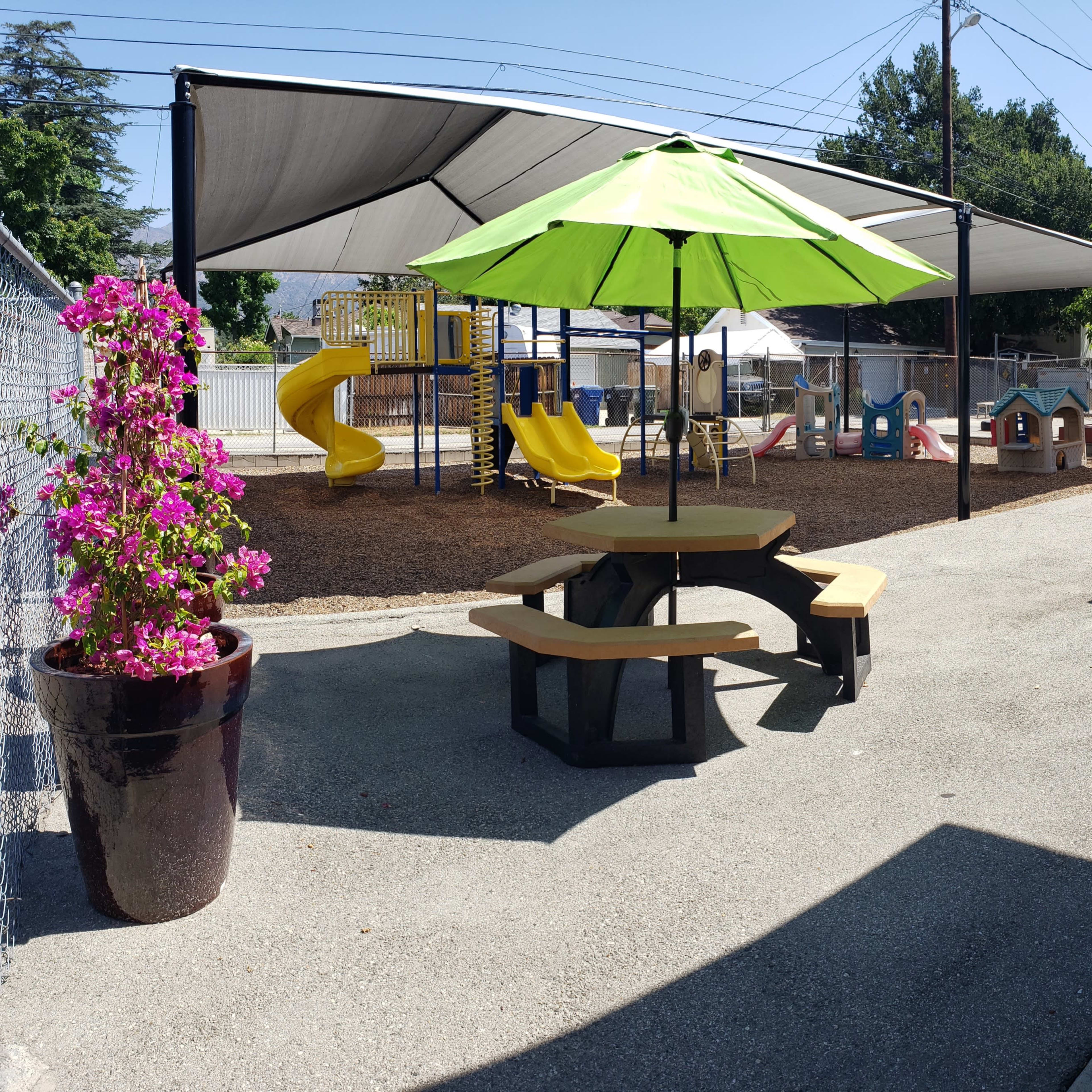 A playground area with a slide, colorful play structures, and two picnic tables shaded by green umbrellas.