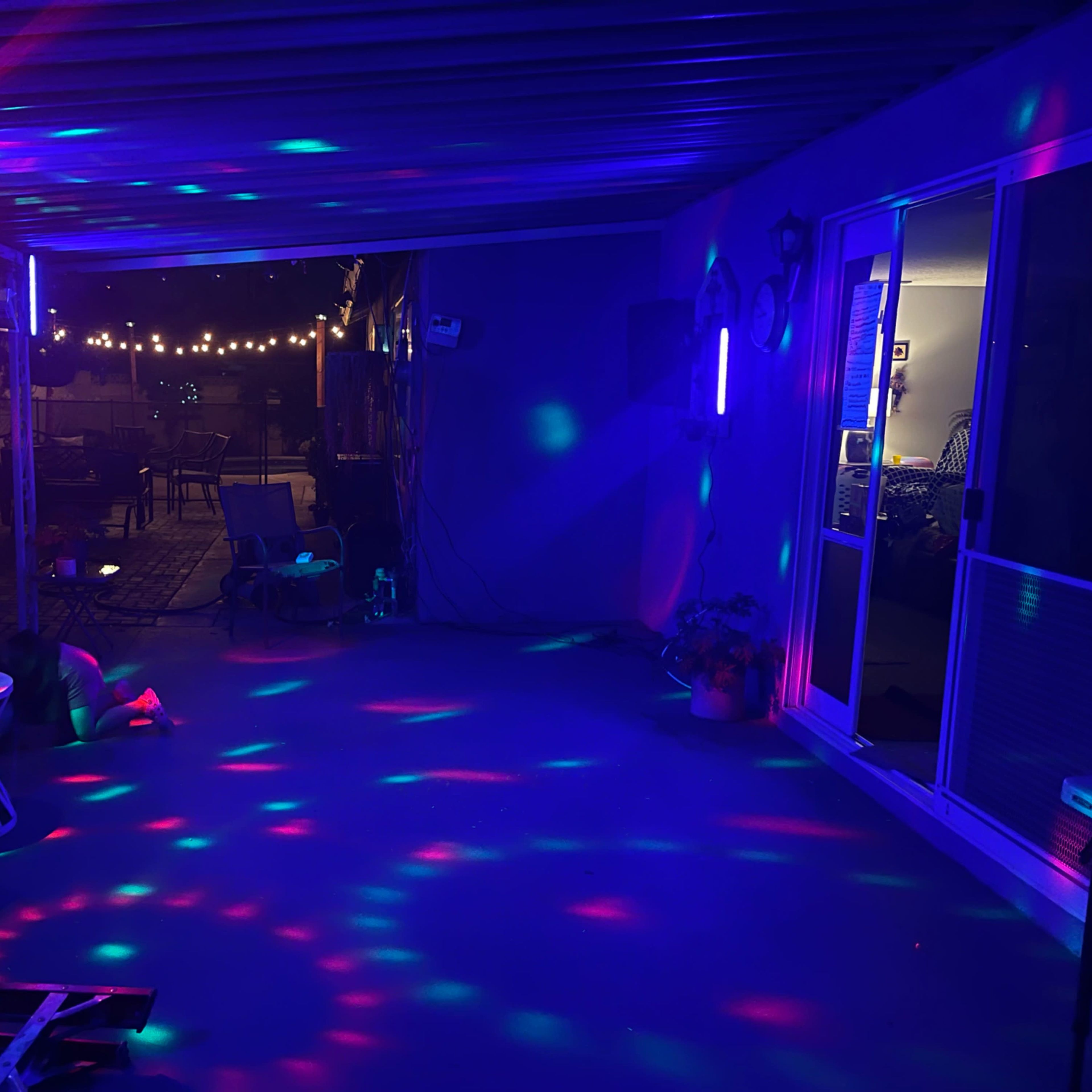 A patio area is illuminated with colorful lights casting patterns on the floor, while string lights hang above in the background.