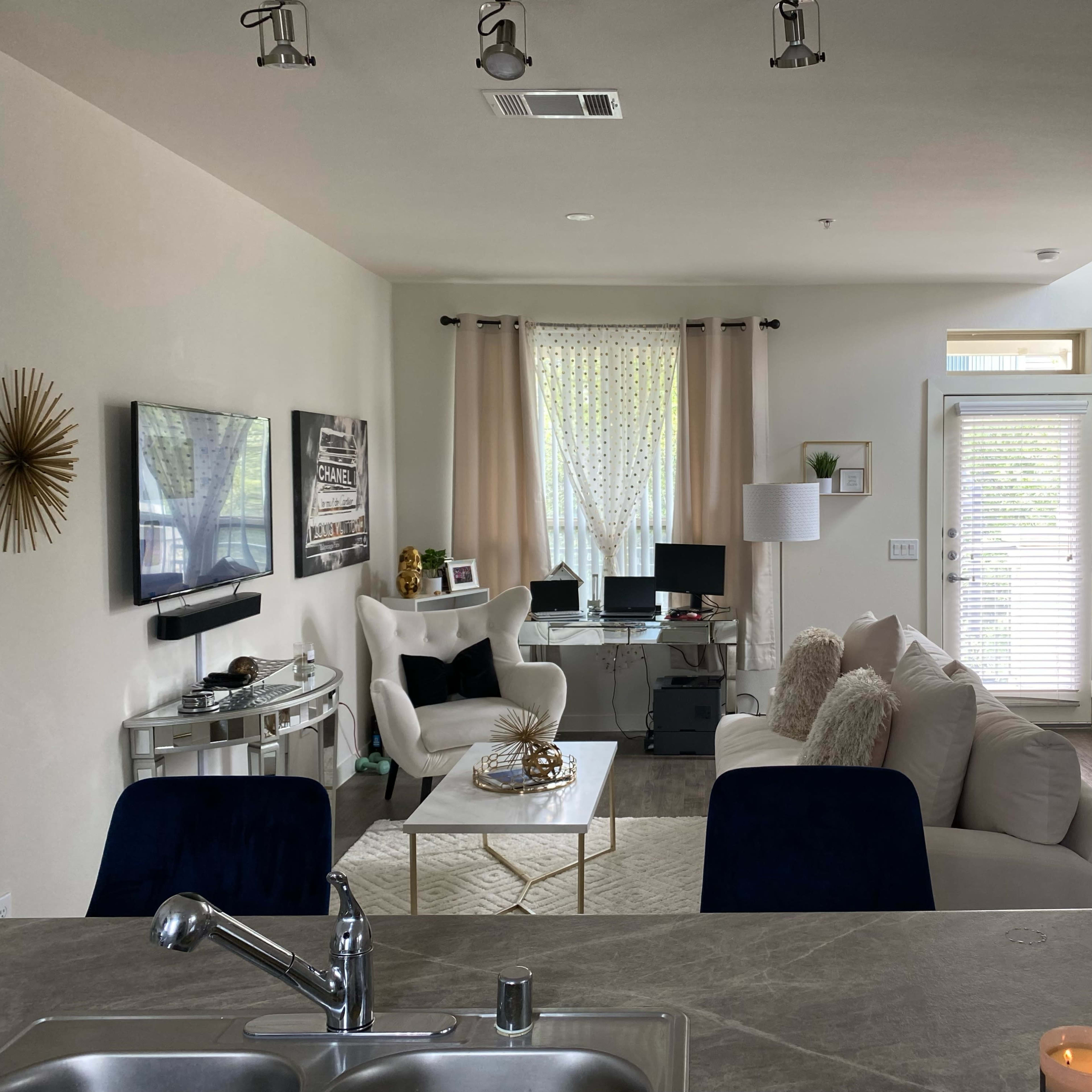 A modern, open-concept living area with a white sofa, a glass coffee table, and a television mounted on the wall, along with large windows letting in natural light.