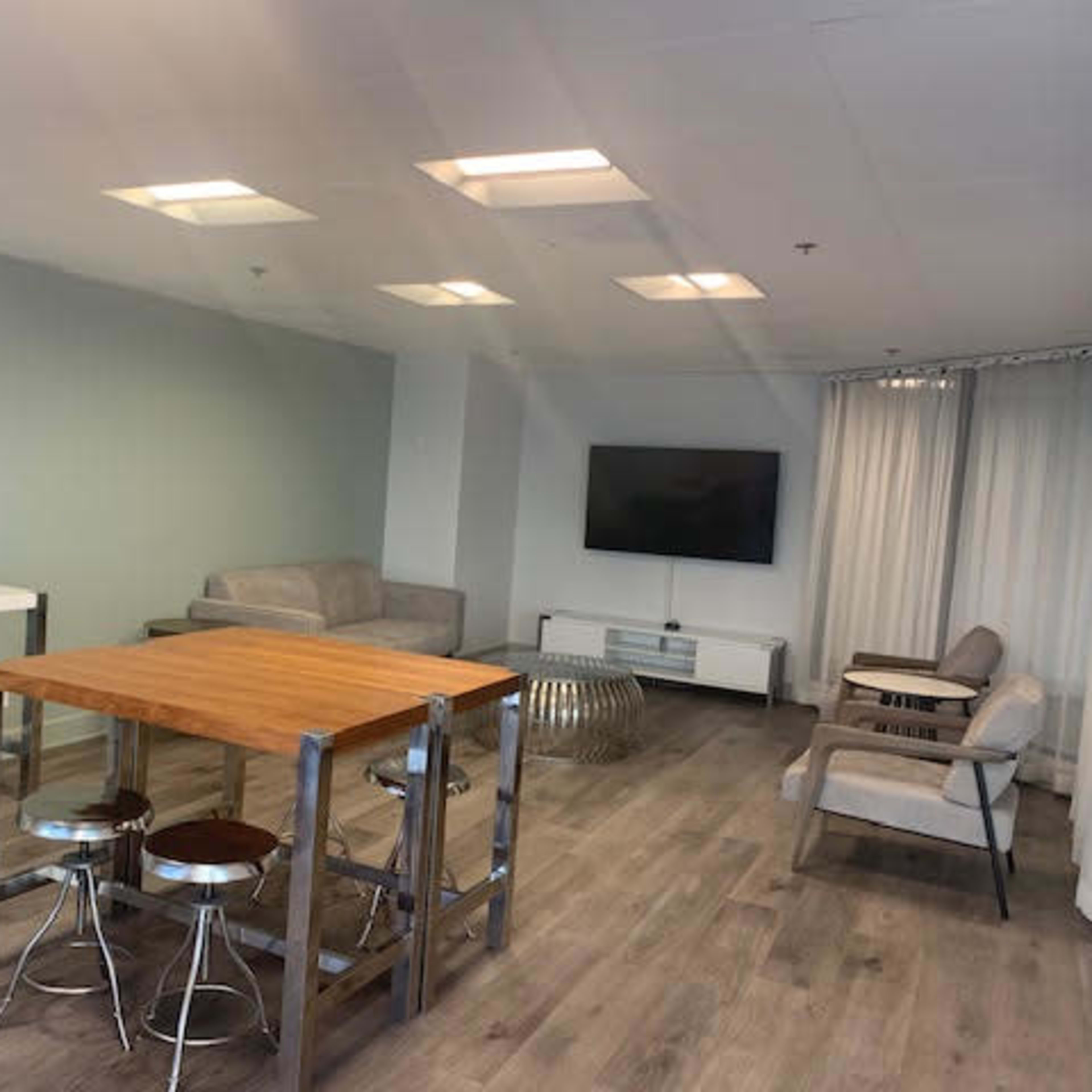 A modern, minimalistic lounge area with a wooden table, metal stools, a sofa, two chairs, and a television mounted on the wall.
