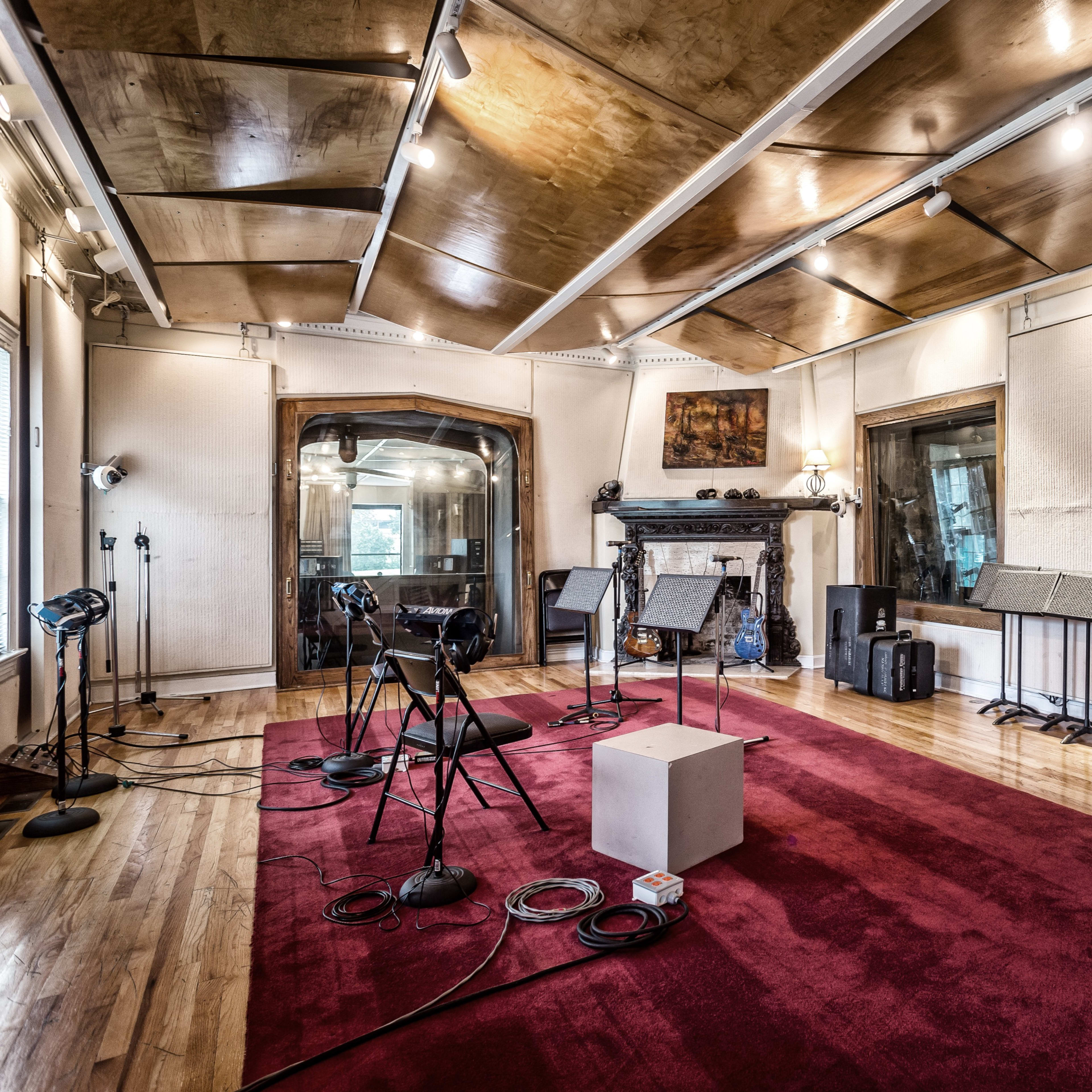A recording studio features several microphones on stands, music stands, and a small white table on a red carpeted floor with wooden paneling and large windows.