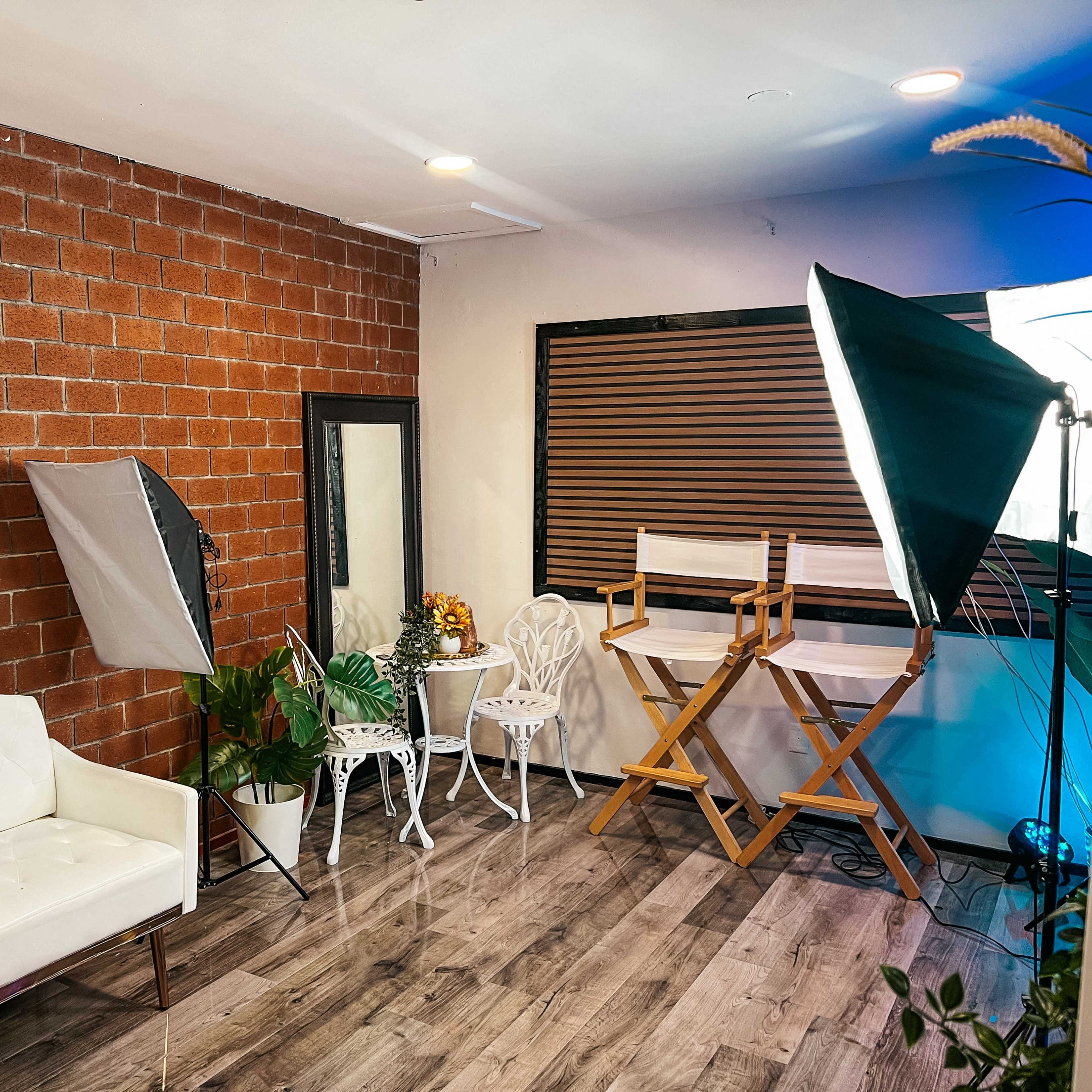 A photography studio features a white couch, a large mirror, two director's chairs, and lighting equipment against a brick wall.