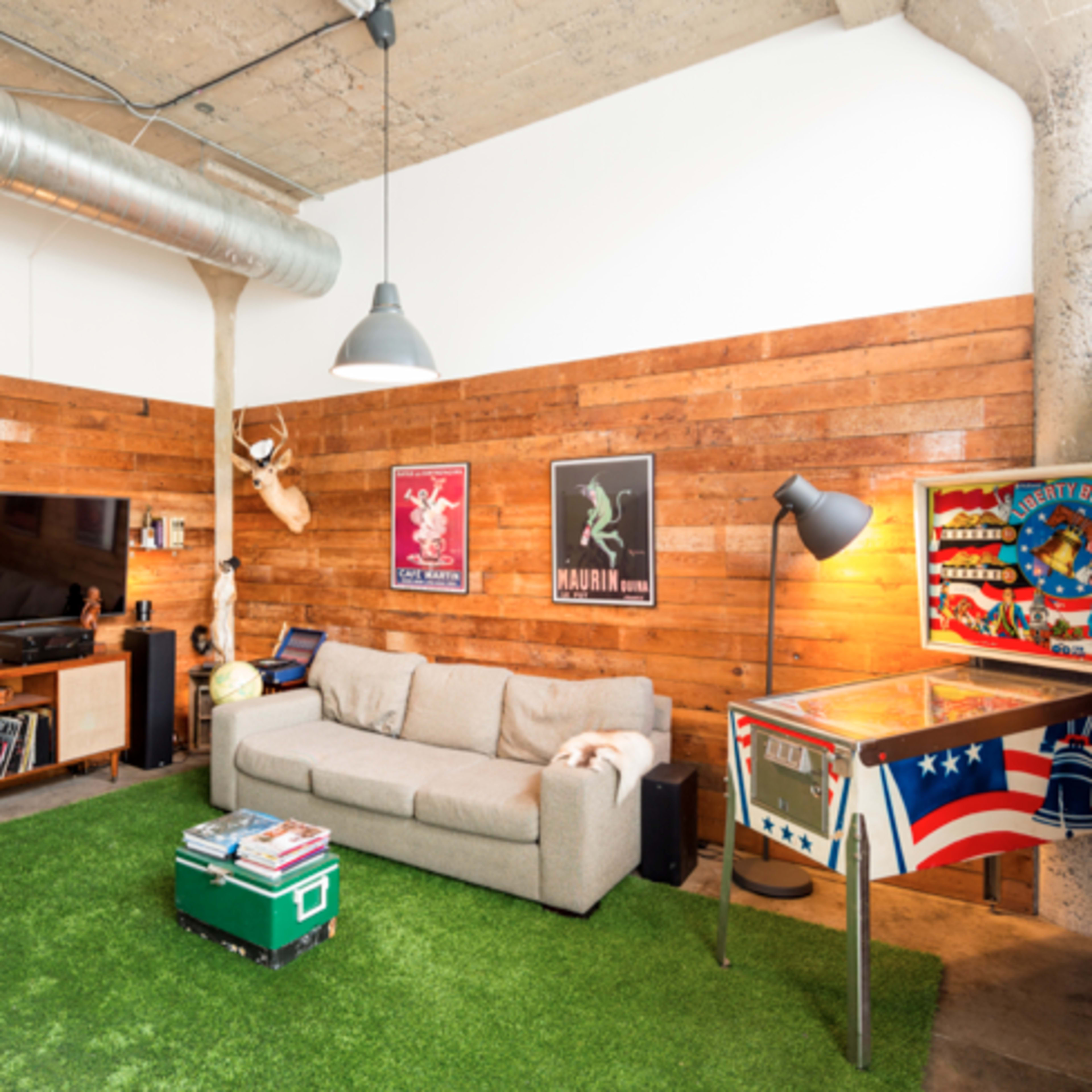 A modern living room with wooden walls, a gray sofa, a TV, a pinball machine, and a green area rug.