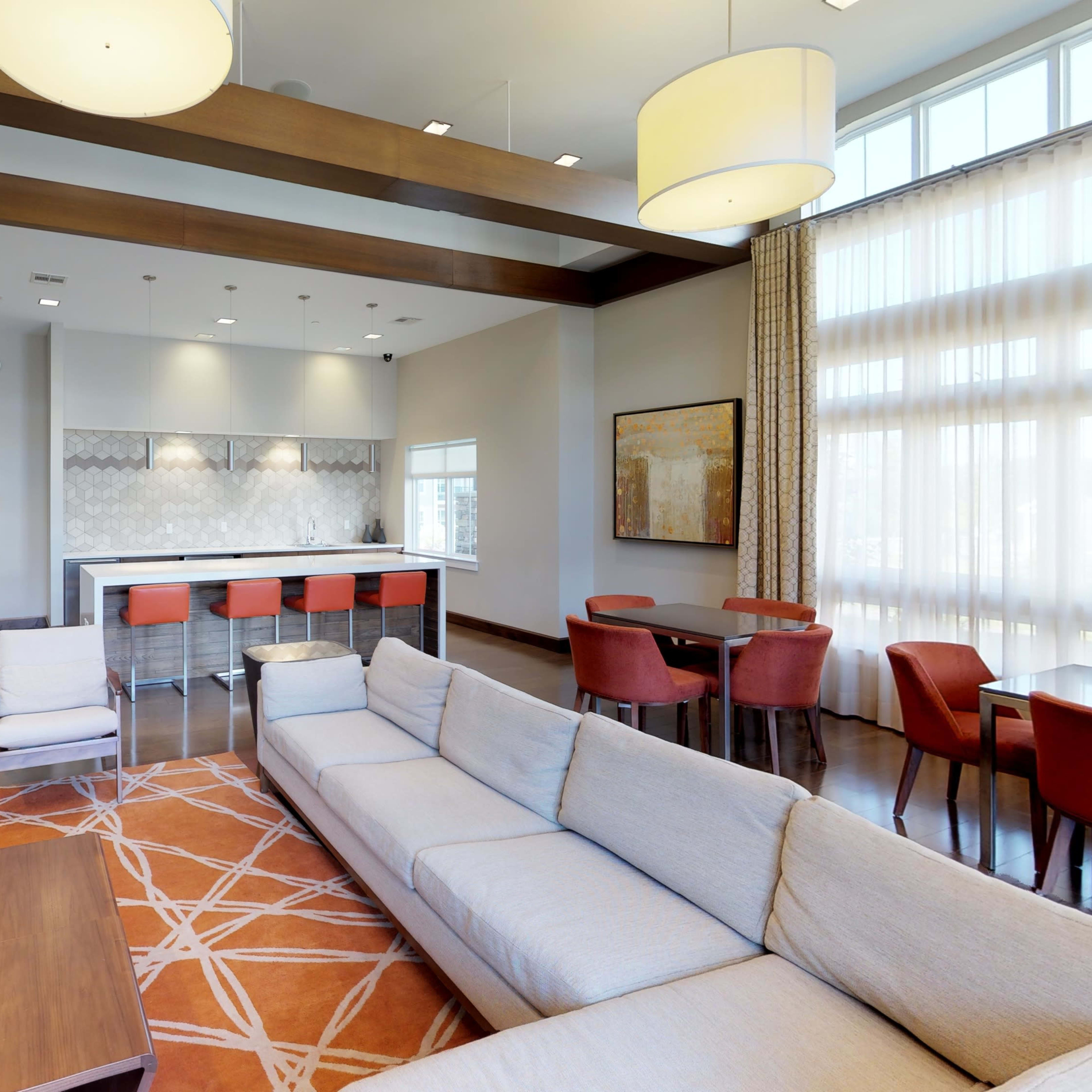 A modern lounge area with a sectional sofa, a coffee table, and a bar with stools, illuminated by pendant lighting and large windows.
