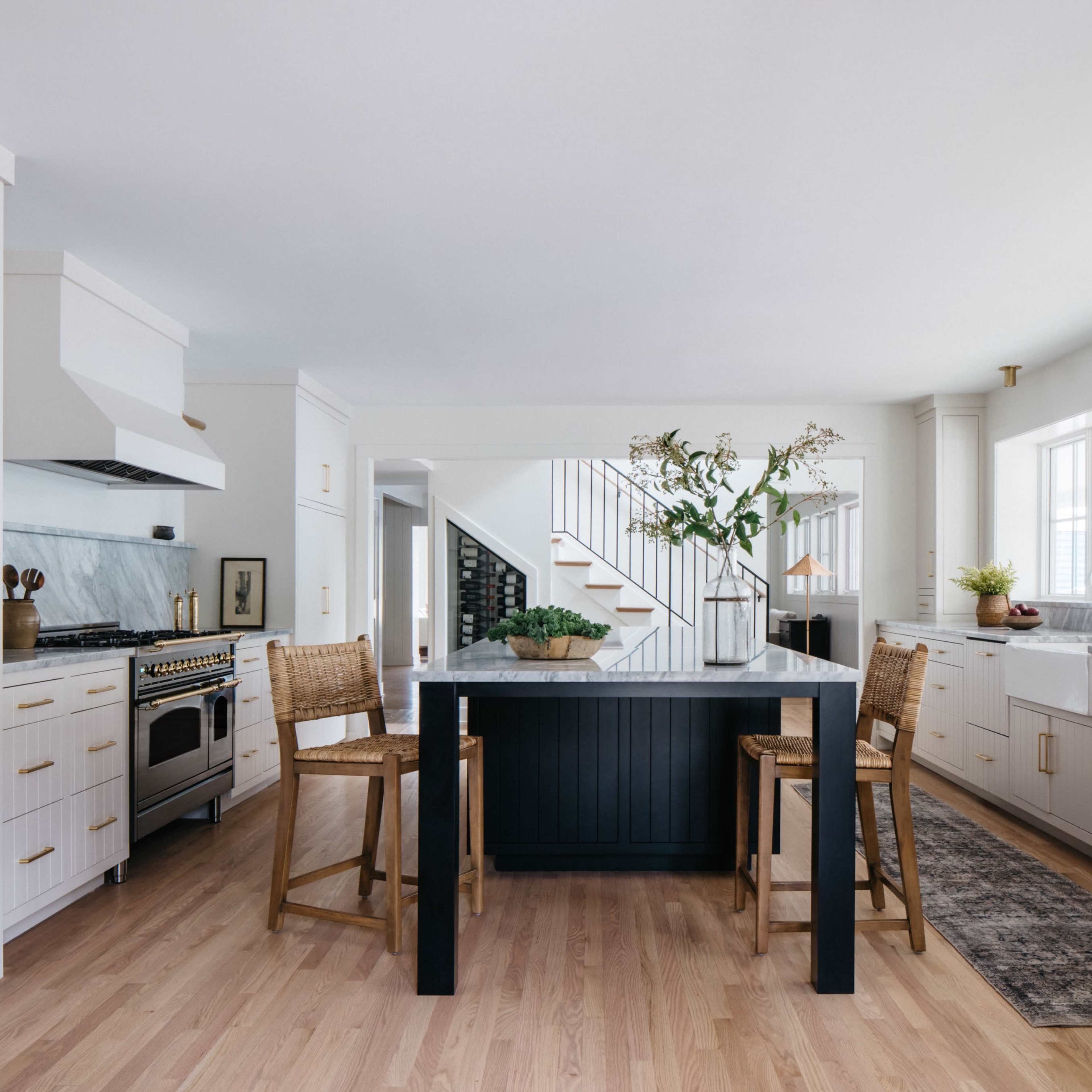 A modern kitchen features a central island with bar stools, white cabinetry, a marble countertop, and large windows allowing natural light.