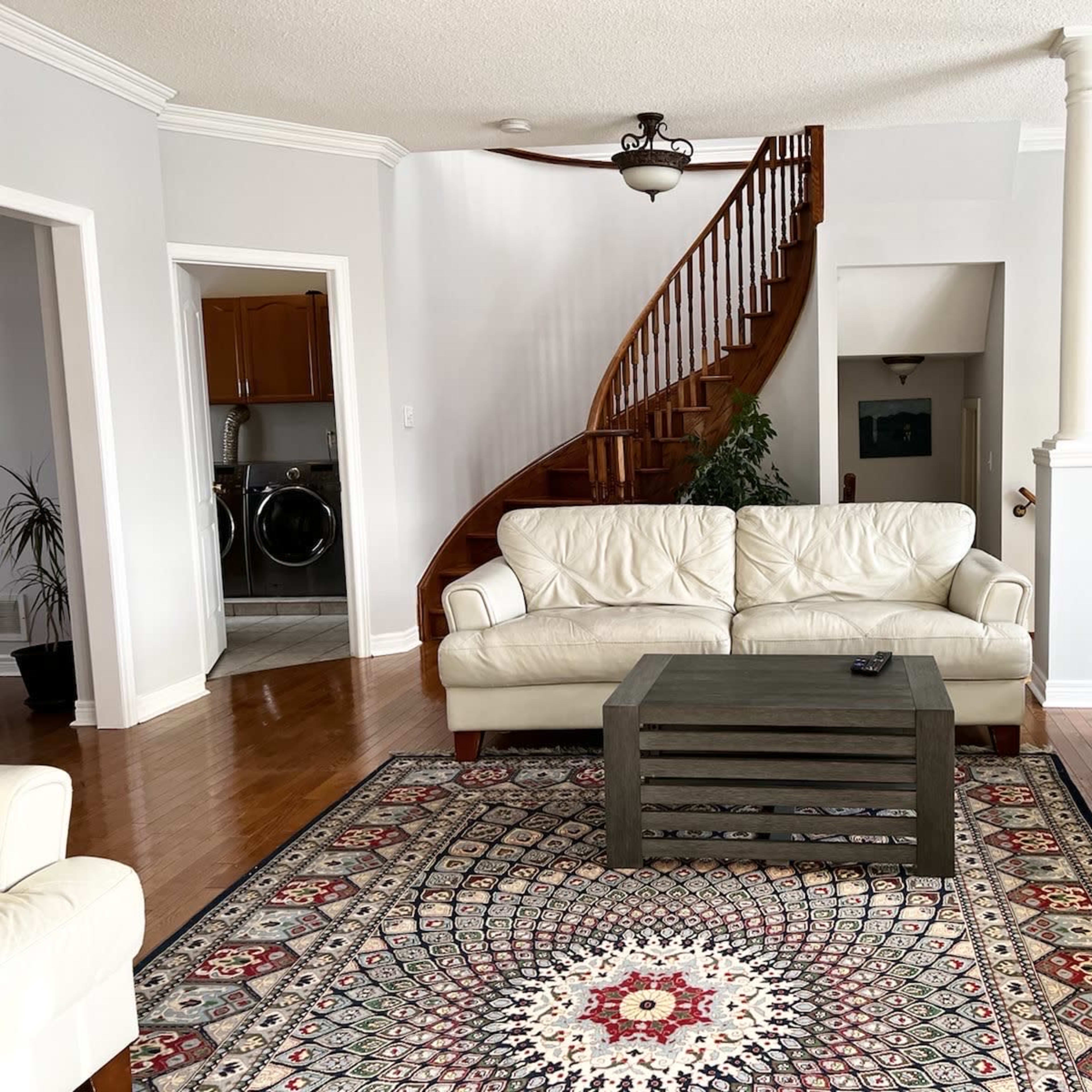 A living room with two white couches facing a wooden coffee table, a patterned area rug on hardwood flooring, and a staircase in the background.