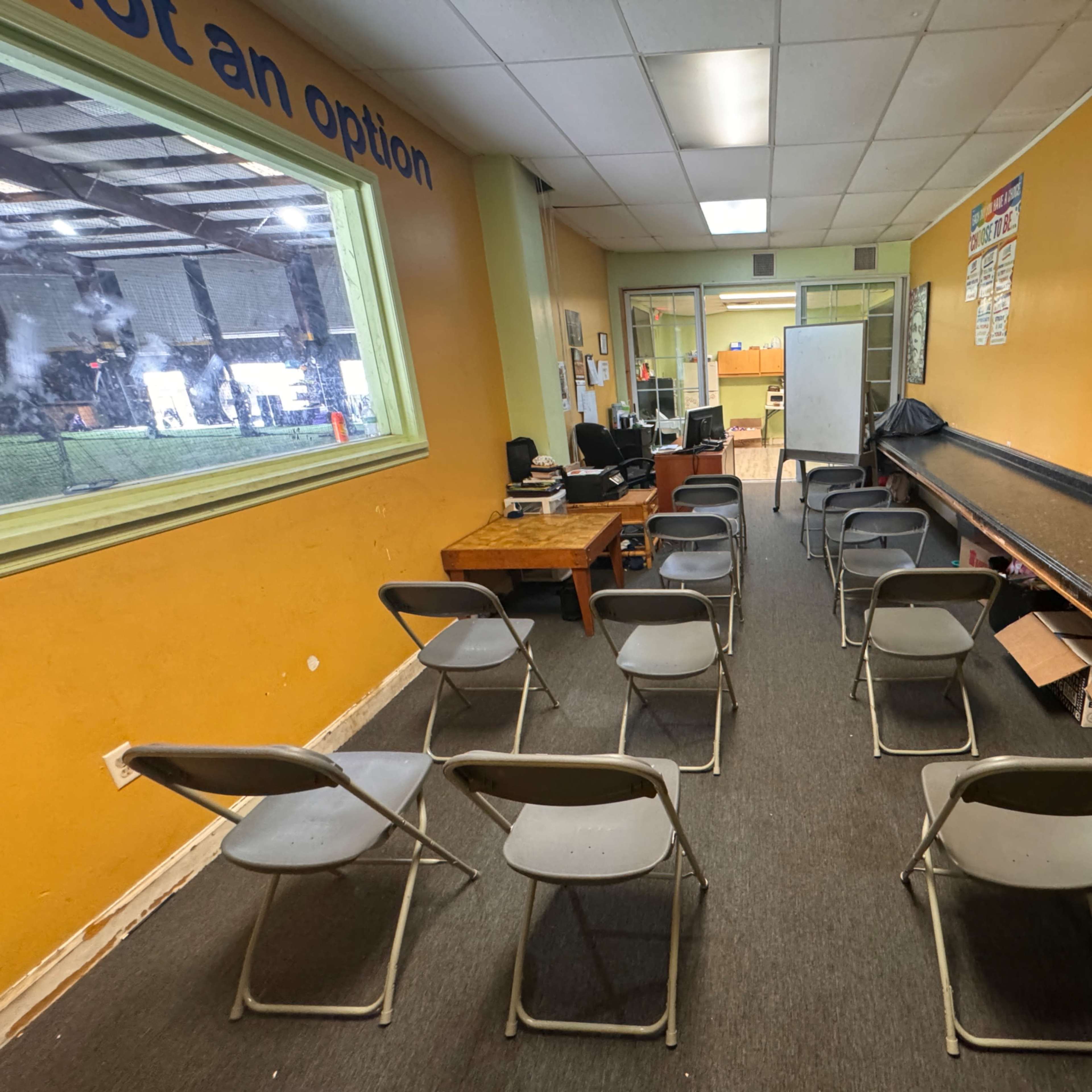 A room with a row of gray folding chairs facing a whiteboard, a desk with equipment in the corner, and a window revealing an outdoor area.