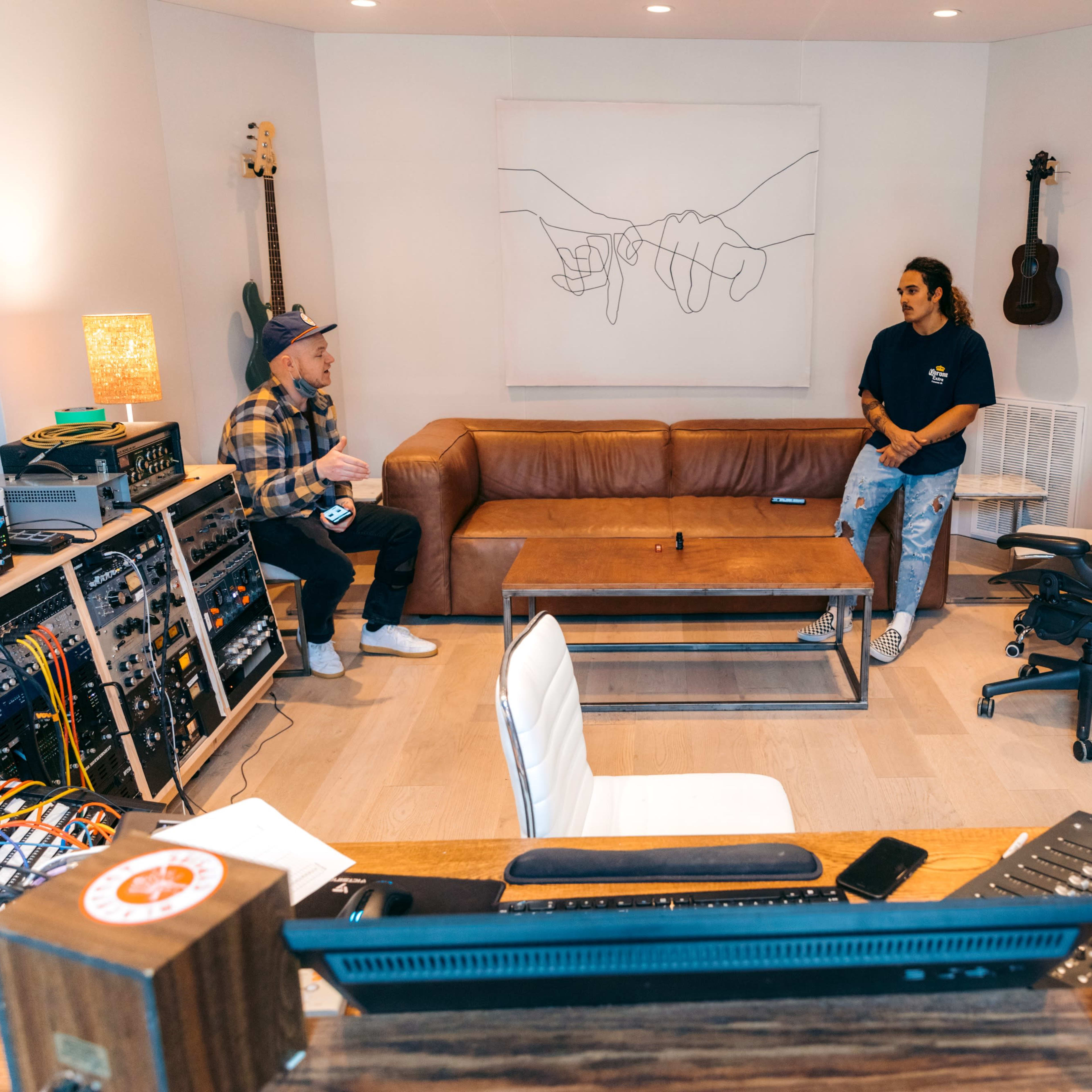 Two people converse in a modern studio space featuring a leather couch, a glass coffee table, and musical equipment.