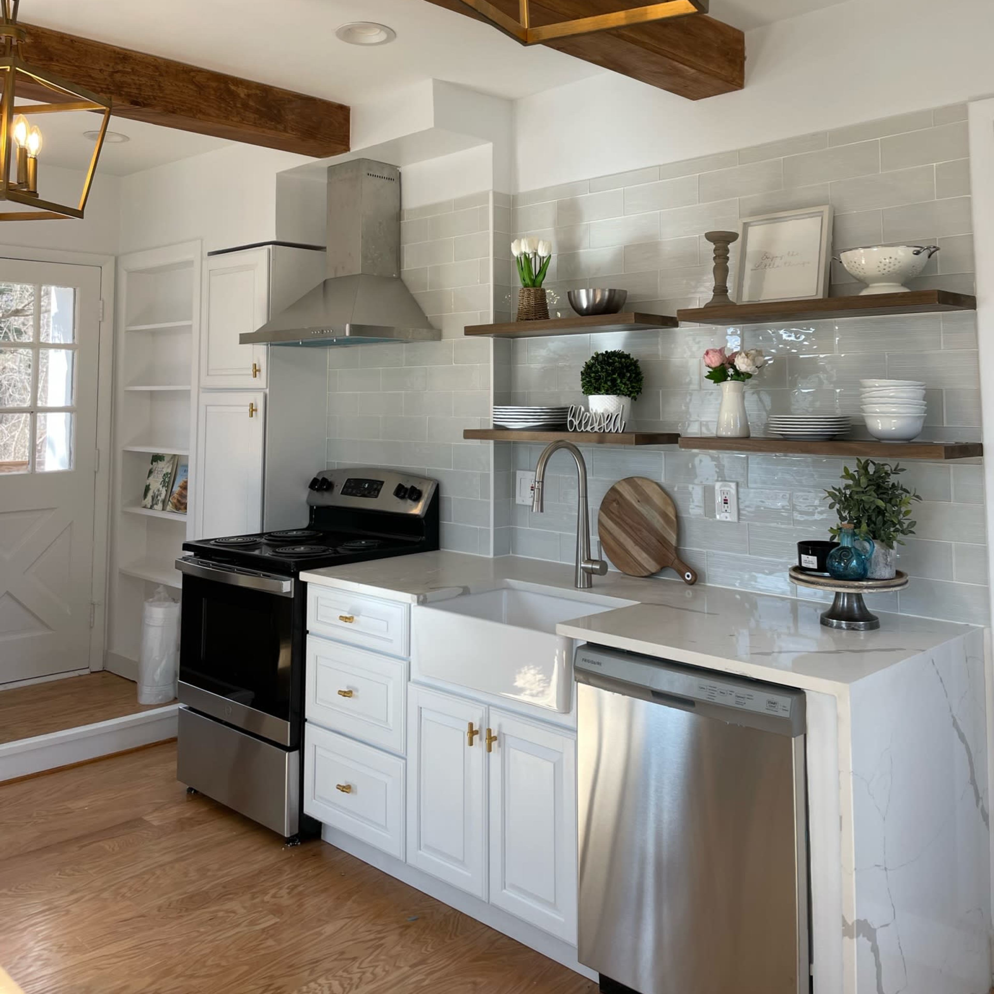 The kitchen features white cabinetry, a stainless steel stove and dishwasher, open shelving, and pendant lighting, with a door leading to another room.