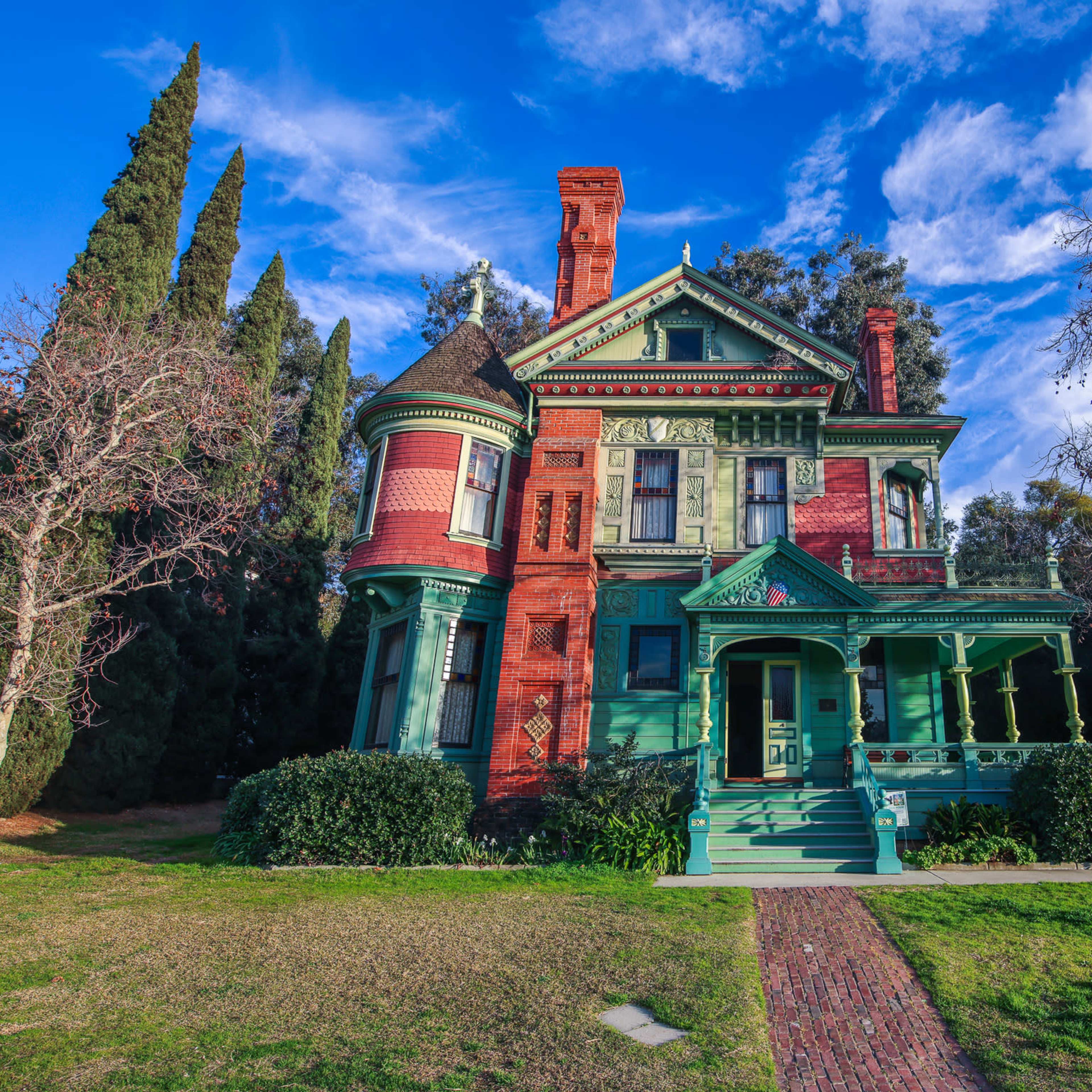 A Victorian-style house features red brick and green trim with ornate architectural details, surrounded by manicured lawn and trees.