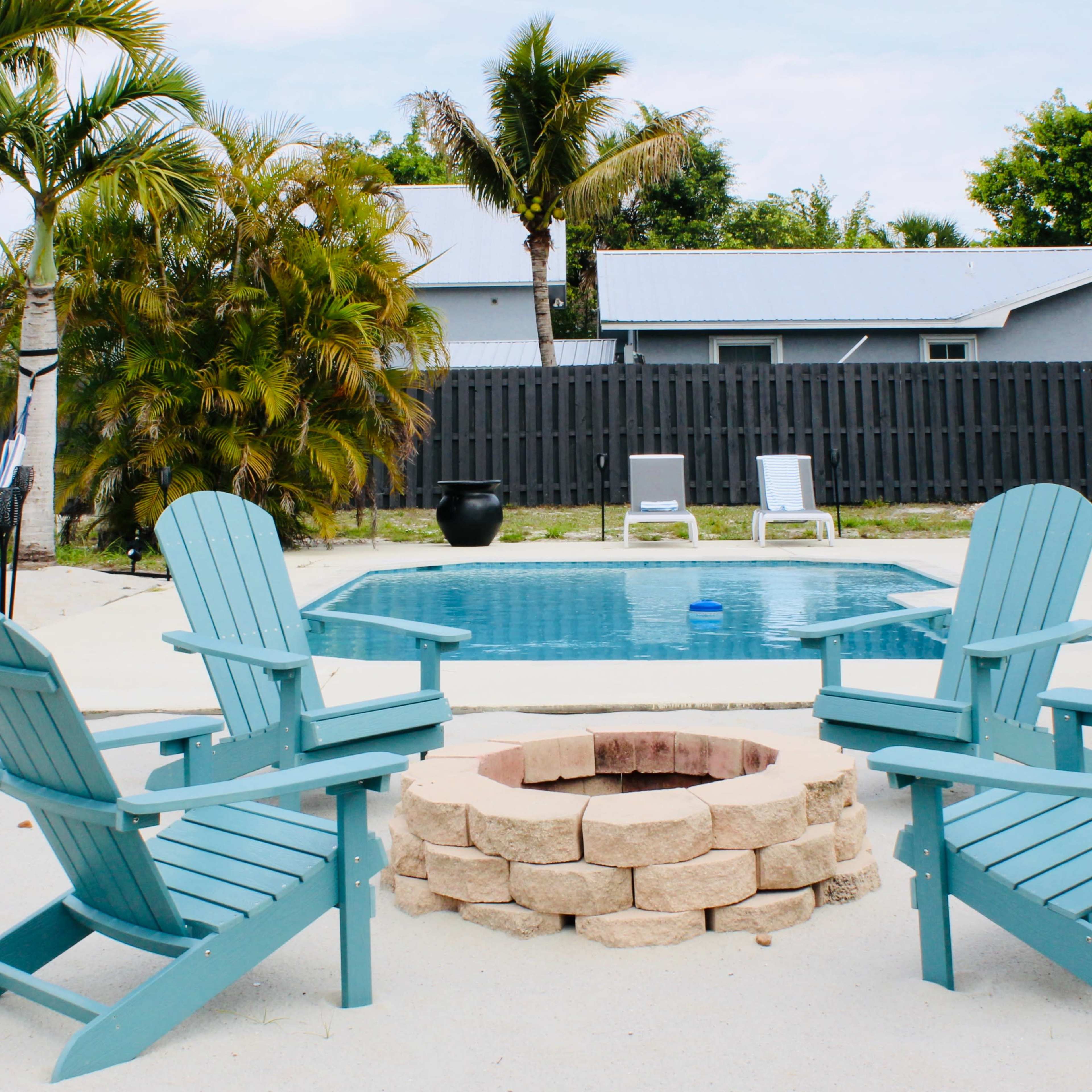 A backyard with a pool surrounded by four blue Adirondack chairs arranged around a stone fire pit, palm trees, and a gray wooden fence in the background.