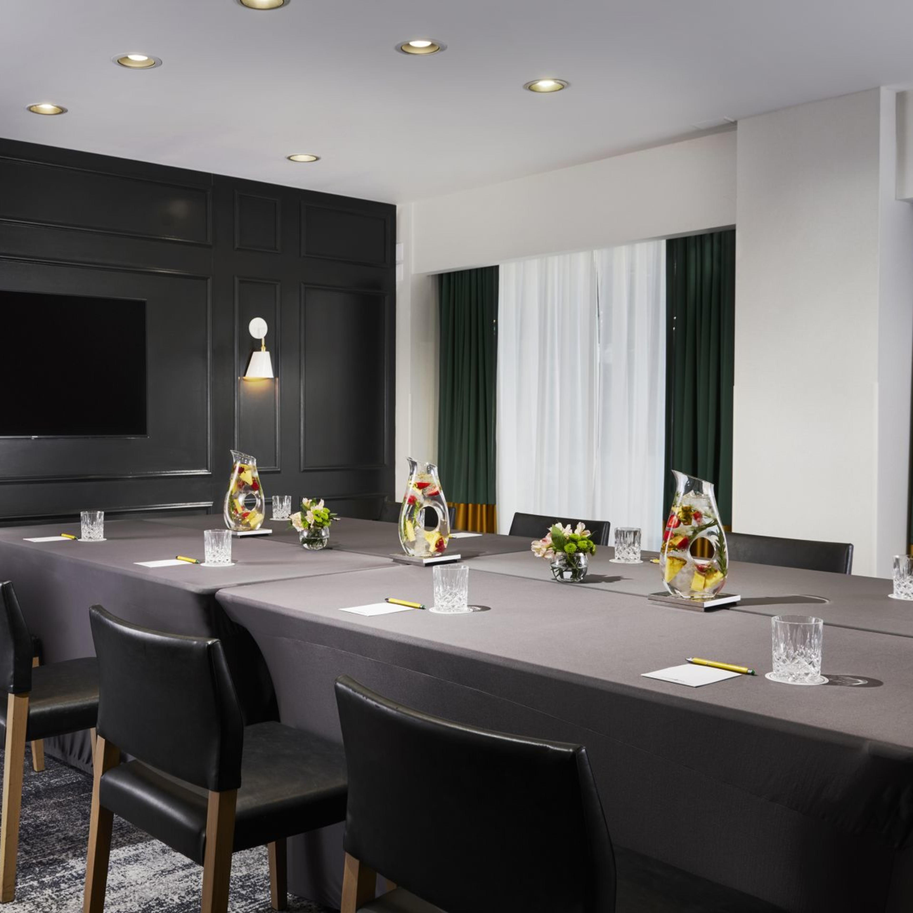A modern conference room with a long dark table, black chairs, and a large screen on the wall, set up for a meeting.