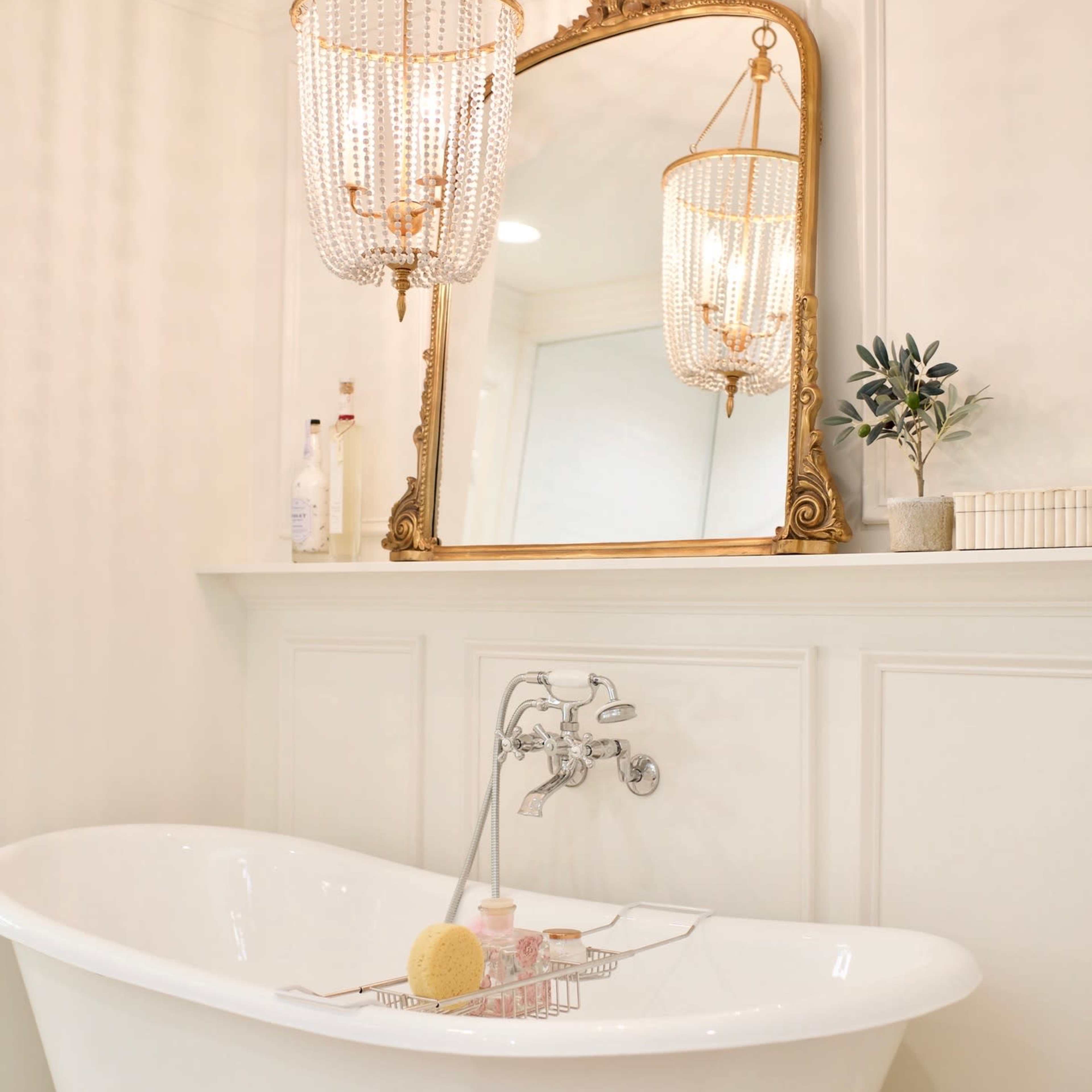 A freestanding white bathtub is displayed under a decorative chandelier and a large ornate mirror in a bright, elegant bathroom.