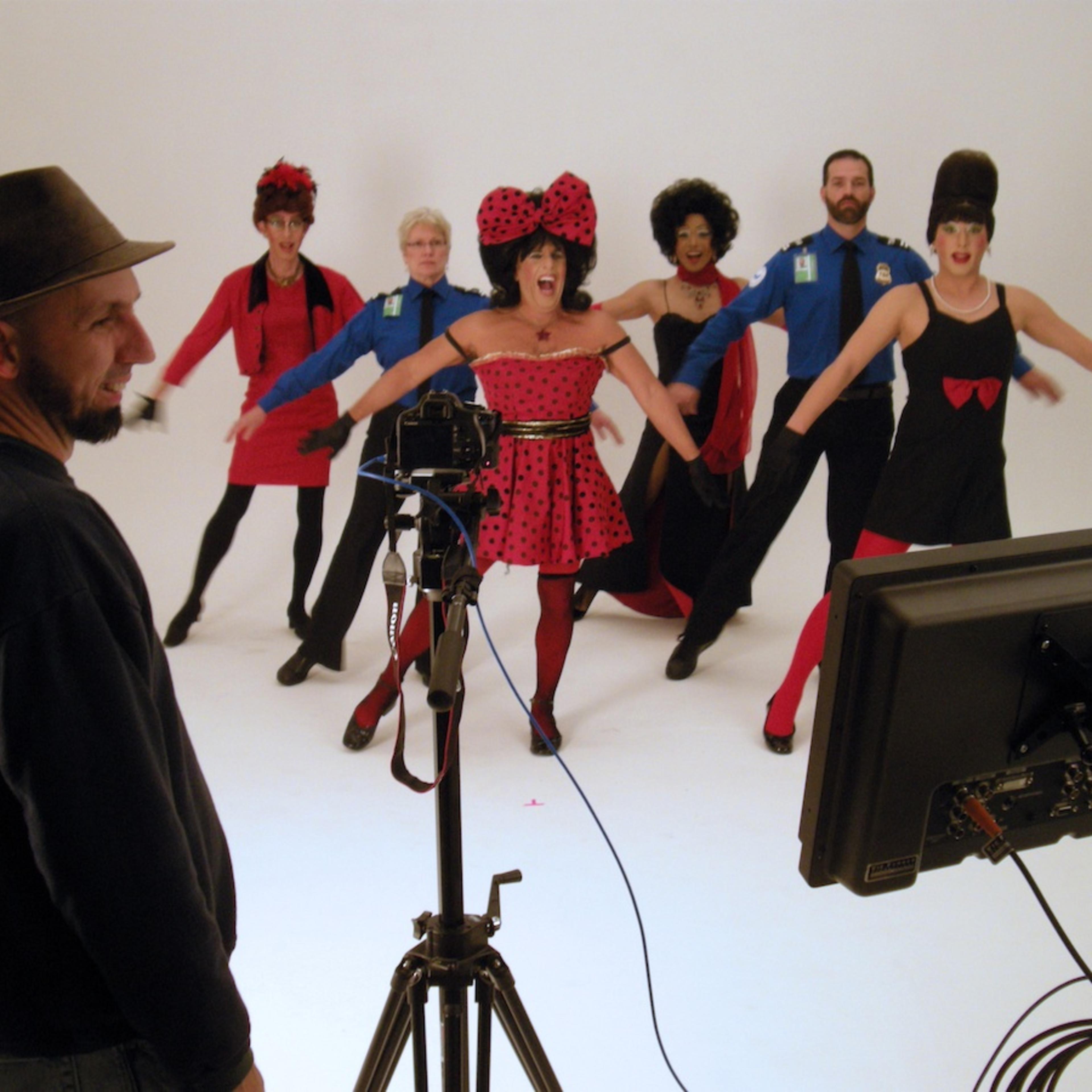 A group of performers in colorful costumes and wigs poses energetically in front of a camera while a man in a hat observes from behind.