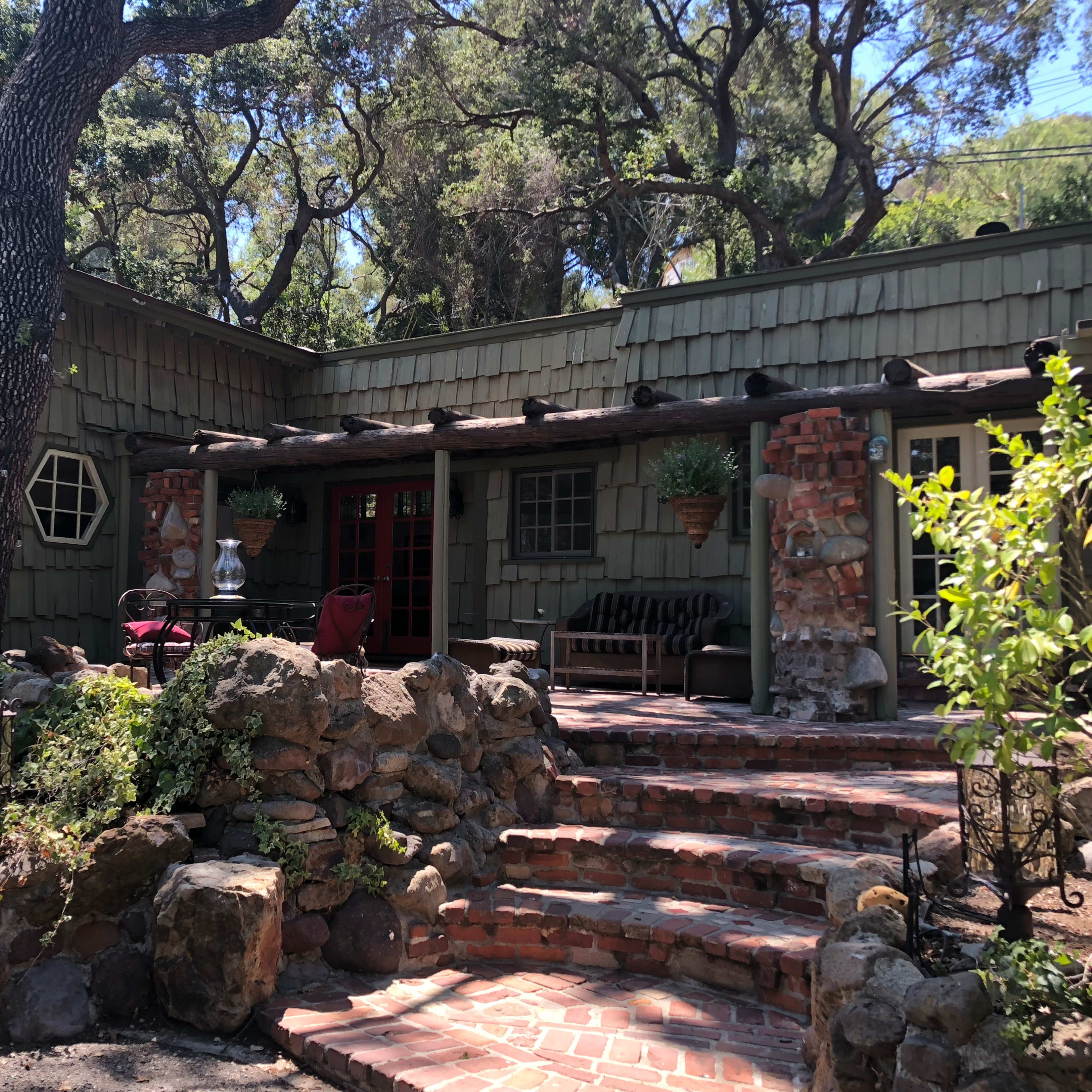 A rustic house with a stone path and a porch, surrounded by greenery and trees.