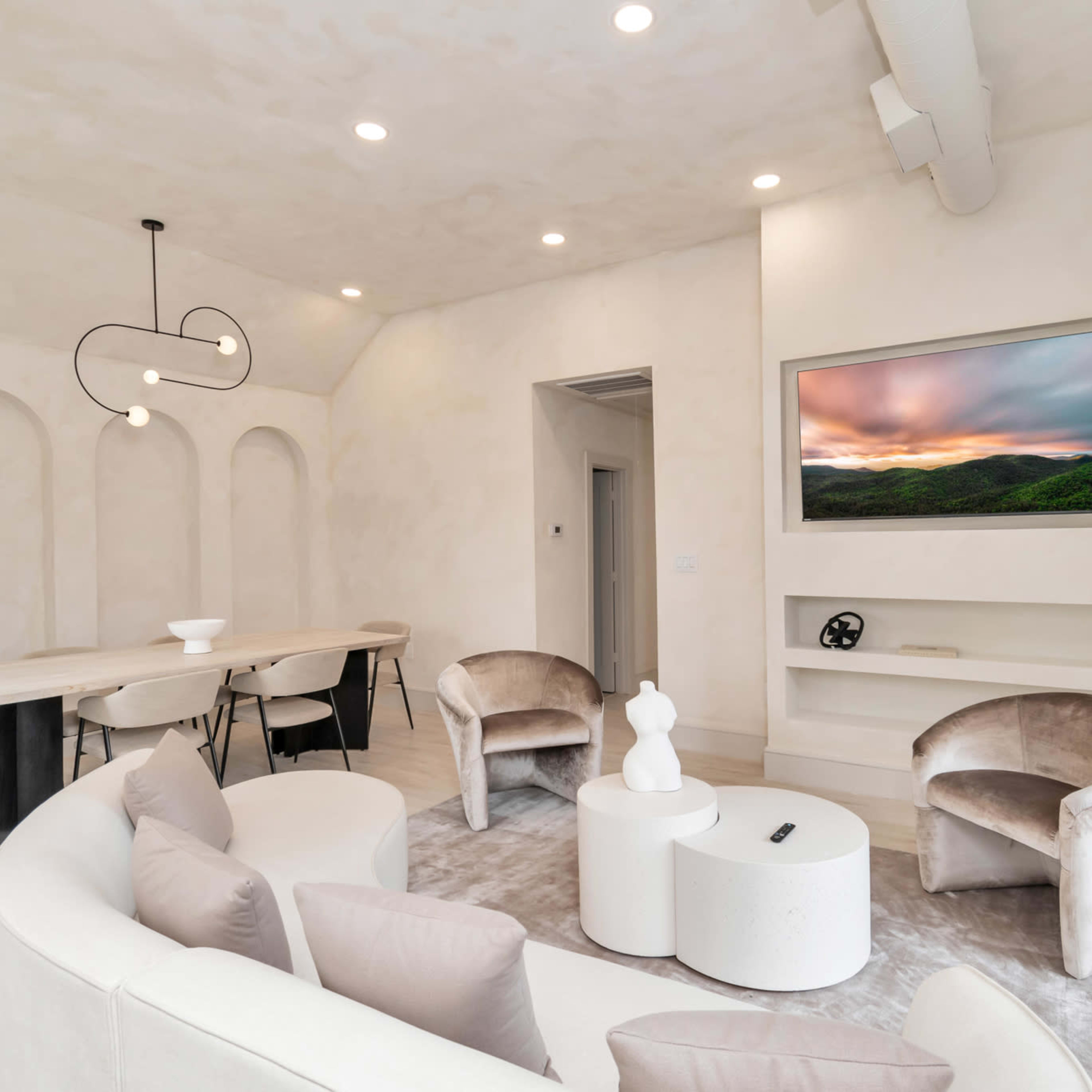 A modern living room with a circular sofa, two armchairs, a coffee table, a dining area with a long table, and a wall-mounted television displaying a landscape.