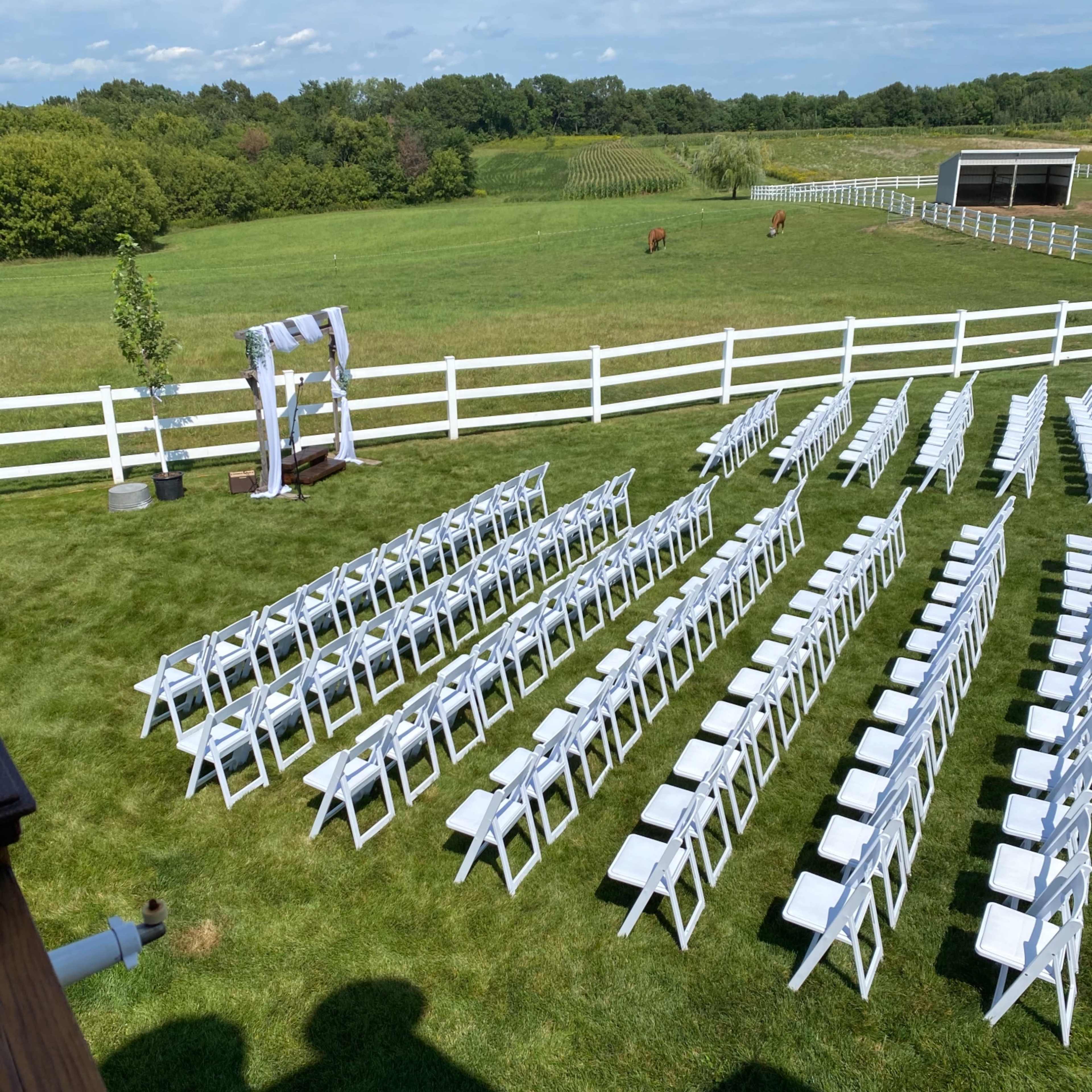 A neatly arranged outdoor wedding setup with white chairs facing an archway on a lawn surrounded by greenery and a white fence.