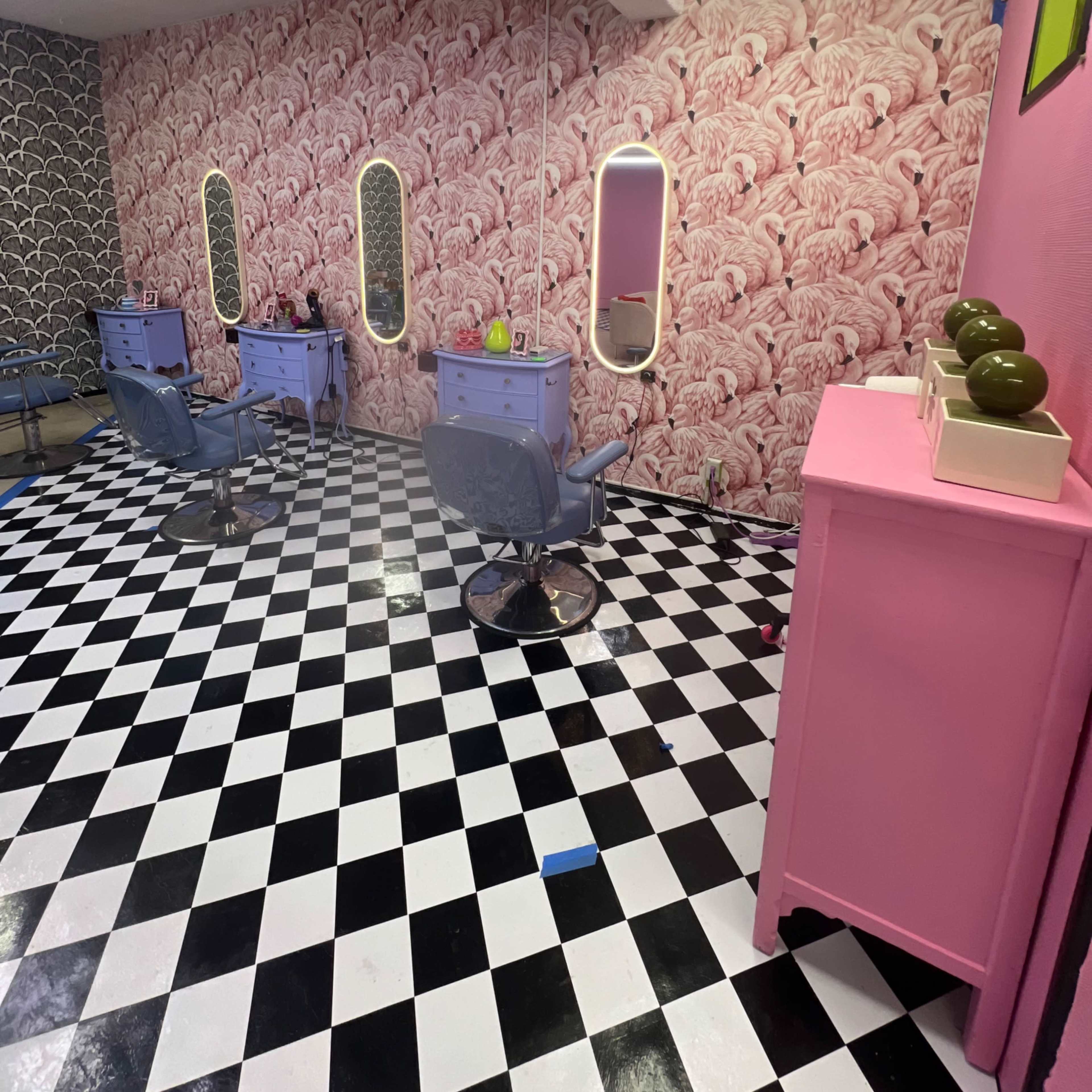 A modern hair salon with pink and black decor, featuring multiple styling stations with mirrors, blue chairs, and a checkered floor.