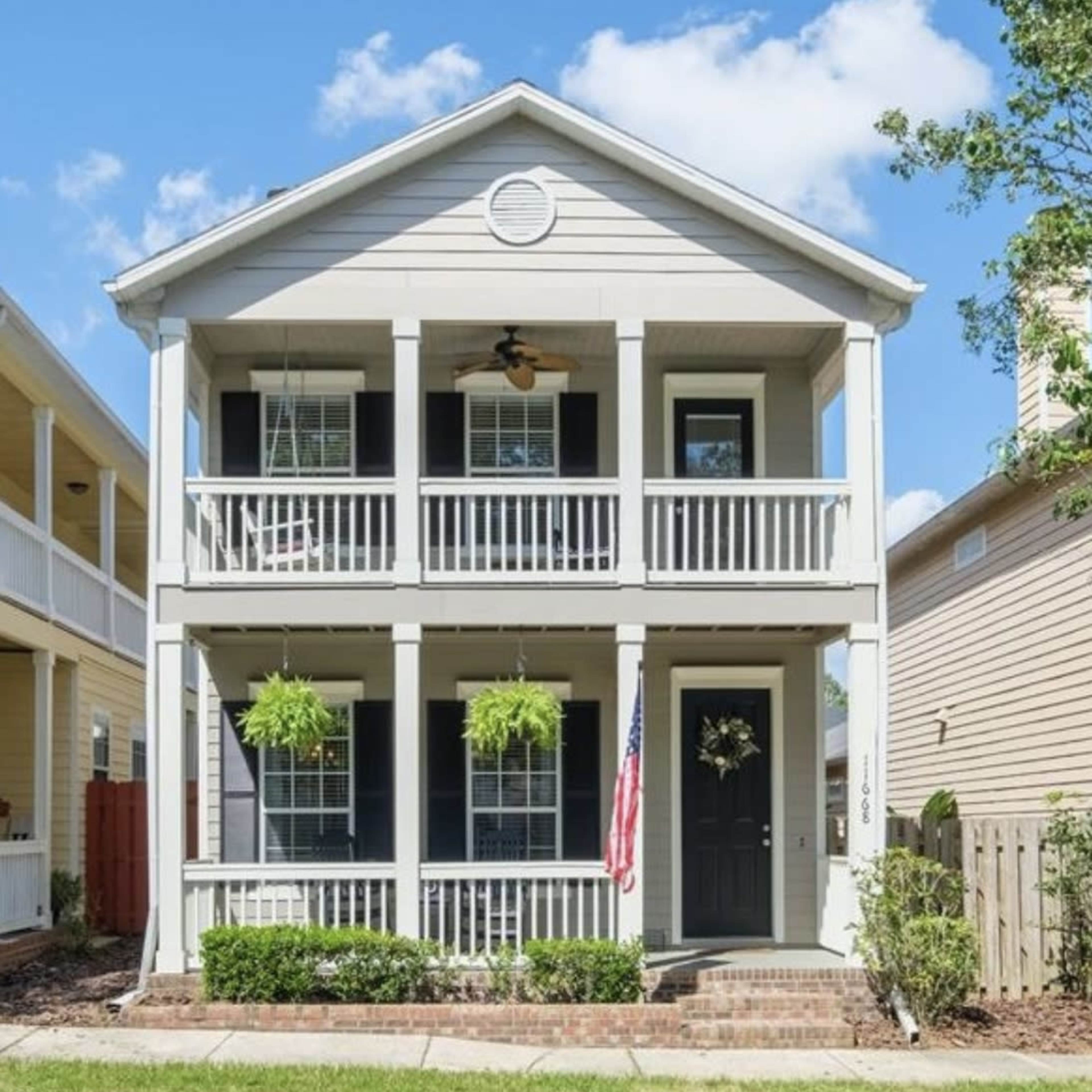 A two-story, light-colored house with a front porch, hanging plants, and an American flag displayed near the entrance.